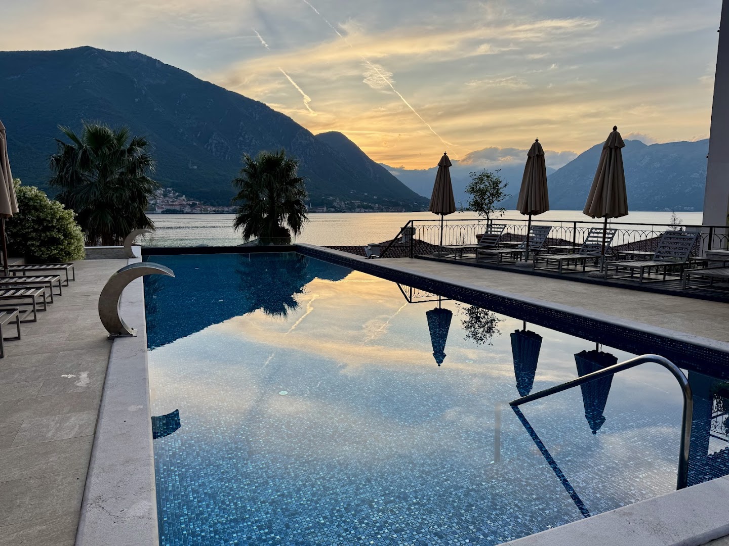 HUMA Kotor Bay Hotel & Villas baseina terase saulrietā, kalnu silueti un debesu atspulgs ūdenī Melnkalnē