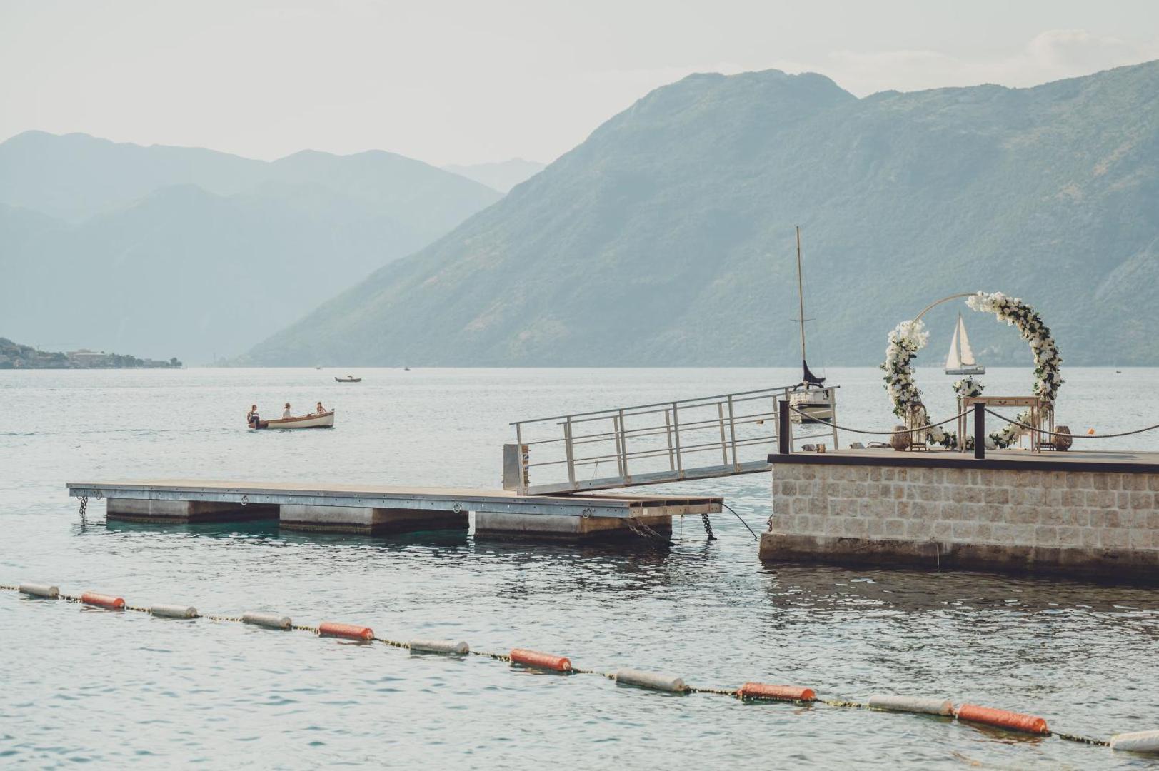 HUMA Kotor Bay Hotel & Villas privātā piestātne ar kāzu ceremonijas arku Kotoras līča piekrastē, Melnkalnē, fonā kalni un laivas