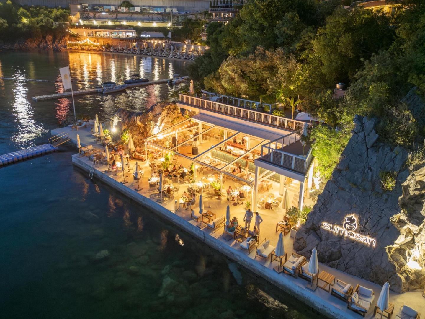 Dukley Hotel & Resort restorāns Sumosan uz platformas pie jūras vakarā — izgaismota terase ar viesiem un skatu uz līci