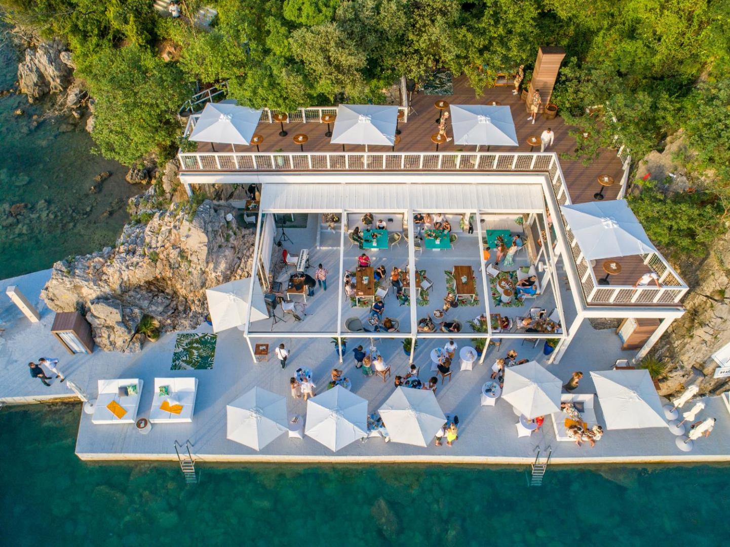 Dukley Hotel & Resort pludmales klubs no augšas dienas laikā — balta terase ar saulessargiem, galdiņiem un piekļuvi tirkīzzilam ūdenim
