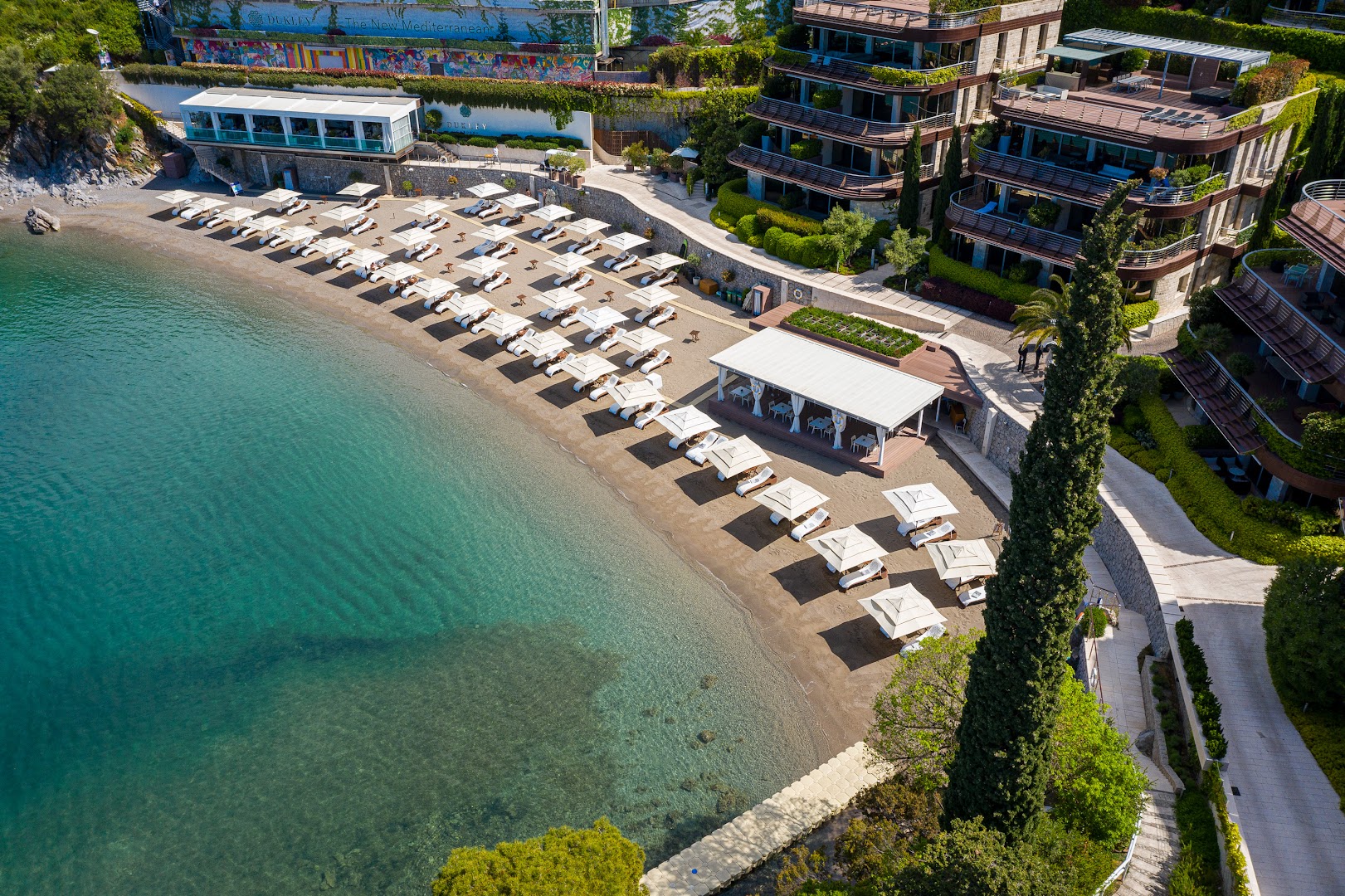 Dukley Hotel & Resort privātā pludmale Budvā, Melnkalnē — skats no augšas uz līci ar saulessargiem, gultām un kristāldzidru ūdeni