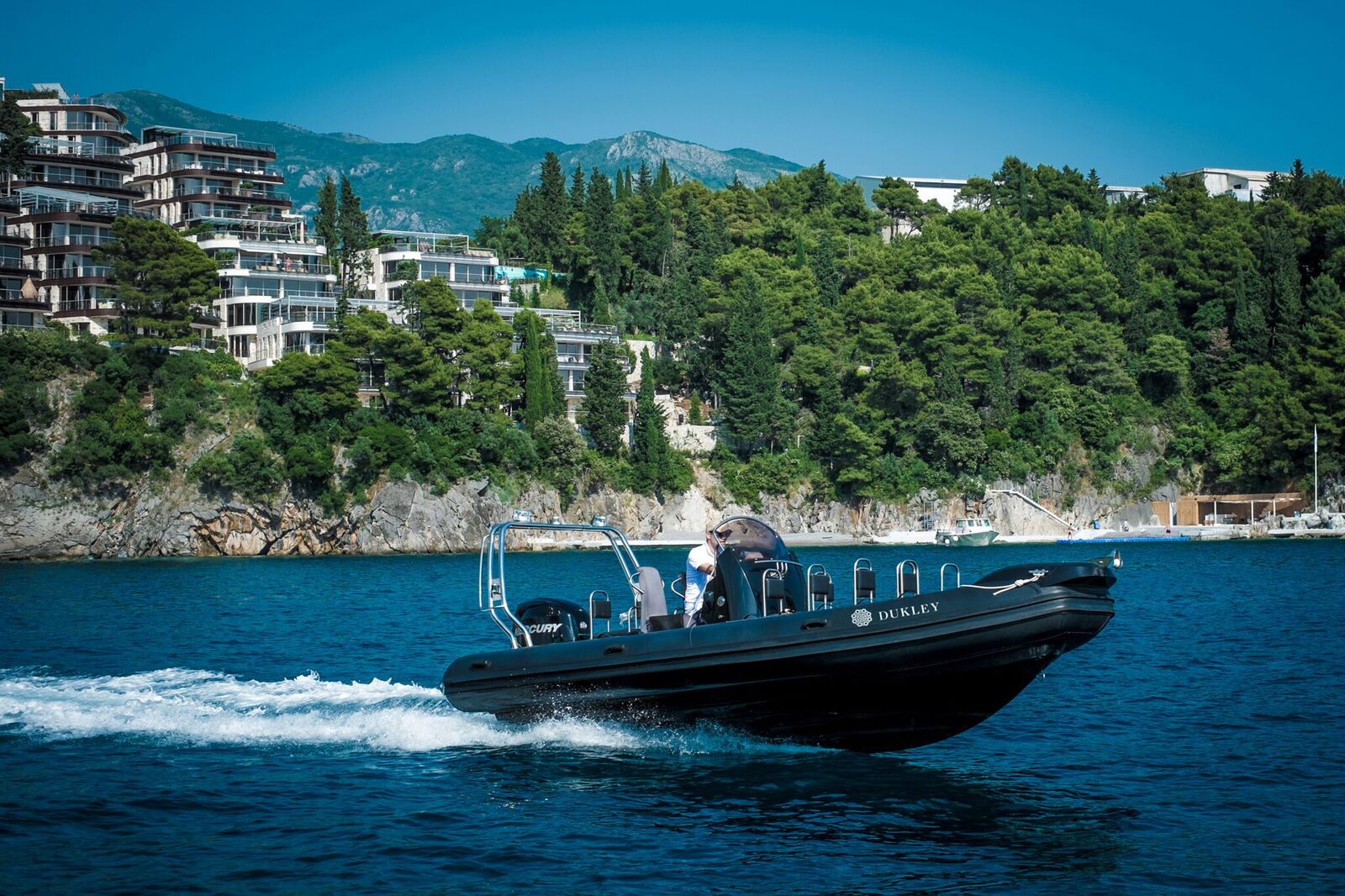 Dukley Hotel & Resort ātrgaitas motorlaiva jūrā uz kalnu un viesnīcas ēku fona — aktīvās atpūtas iespējas Melnkalnē