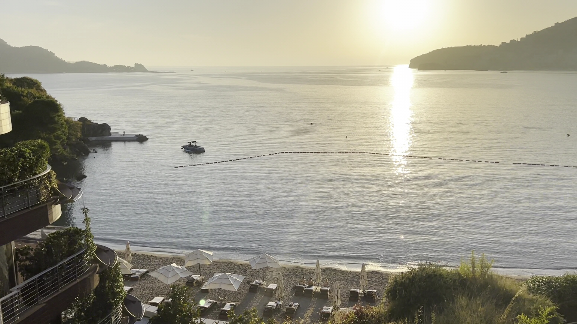 Skats no augšas uz Dukley Hotel & Resort privāto pludmali saulrietā — saulessargi un gultas smilšainajā piekrastē pie rāma ūdens