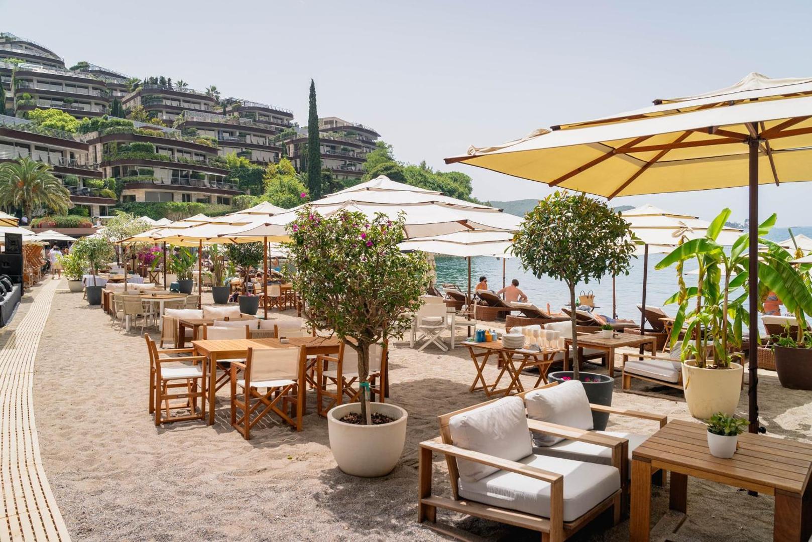 Dukley Hotel & Resort pludmales restorāns ar koka galdiem, baltiem saulessargiem un skatu uz viesnīcas terasveida korpusiem kalna nogāzē