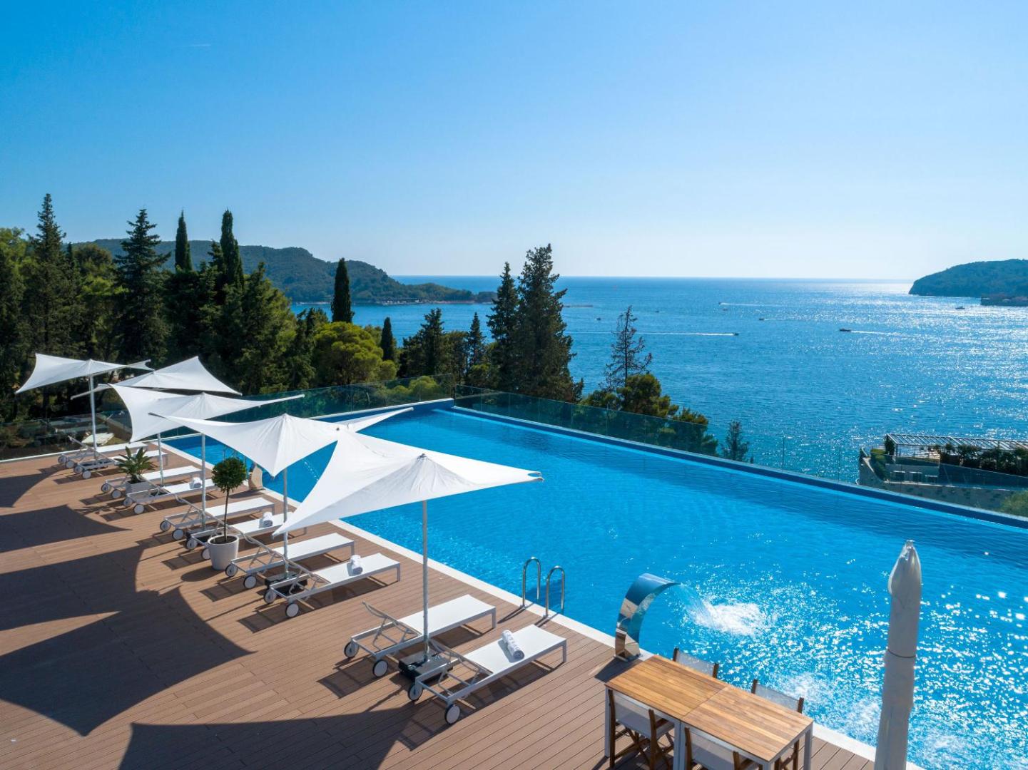Dukley Hotel & Resort bezgalības baseins ar skatu uz Adrijas jūru Budvā — sauļošanās terase ar baltiem saulessargiem un atpūtas krēsliem