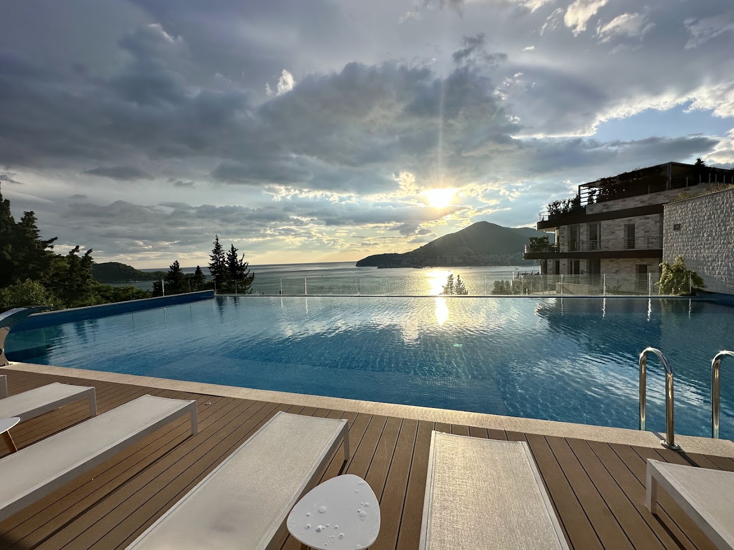 Dukley Hotel & Resort āra baseins saulrieta laikā ar skatu uz kalniem un Budvas līci — atpūtas zona ar moderniem krēsliem