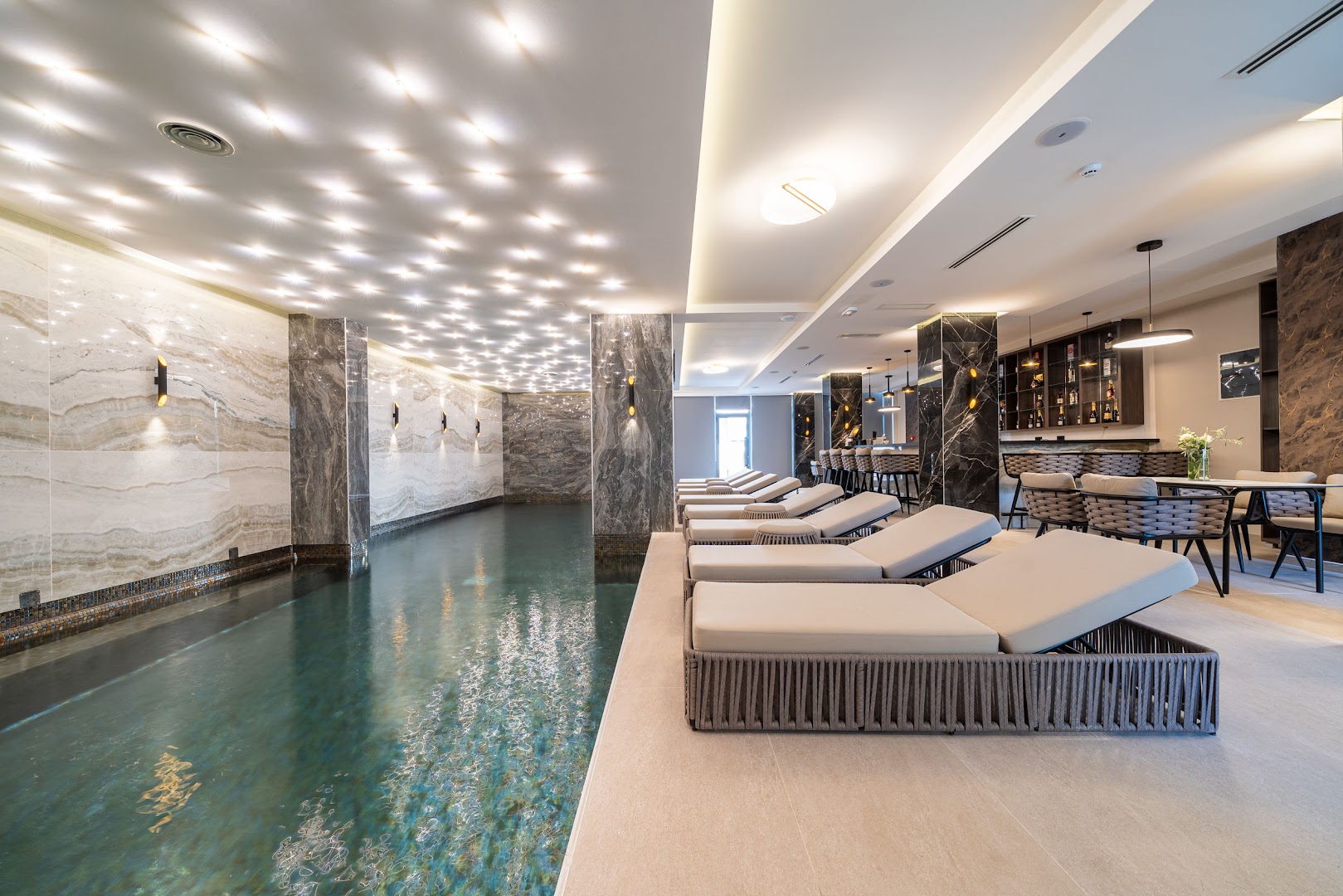 Hotel Millennium by Aycon plašs skats uz iekštelpu baseinu, bāra zonu un atpūtas krēsliem modernā viesnīcas spa kompleksā