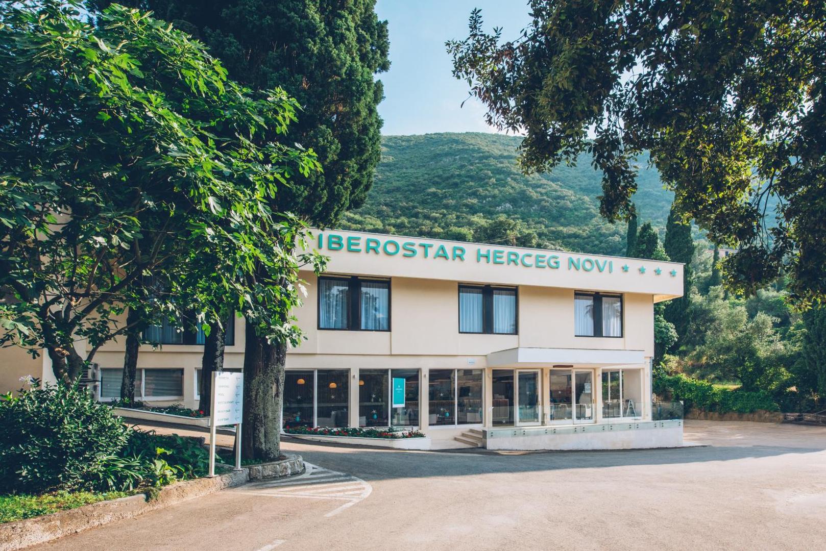 Iberostar Herceg Novi 4* viesnīcas galvenā ieeja un fasāde, ko ieskauj zaļi koki un kalnu ainava Melnkalnē