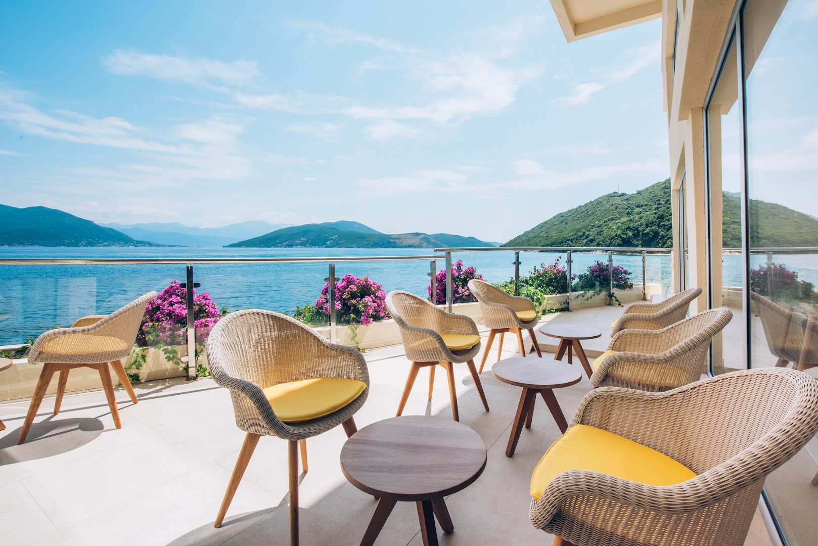 Iberostar Herceg Novi āra terase ar pītiem krēsliem un skatu uz Kotoras līci, kalniem un ziedošiem bugenvillu krūmiem