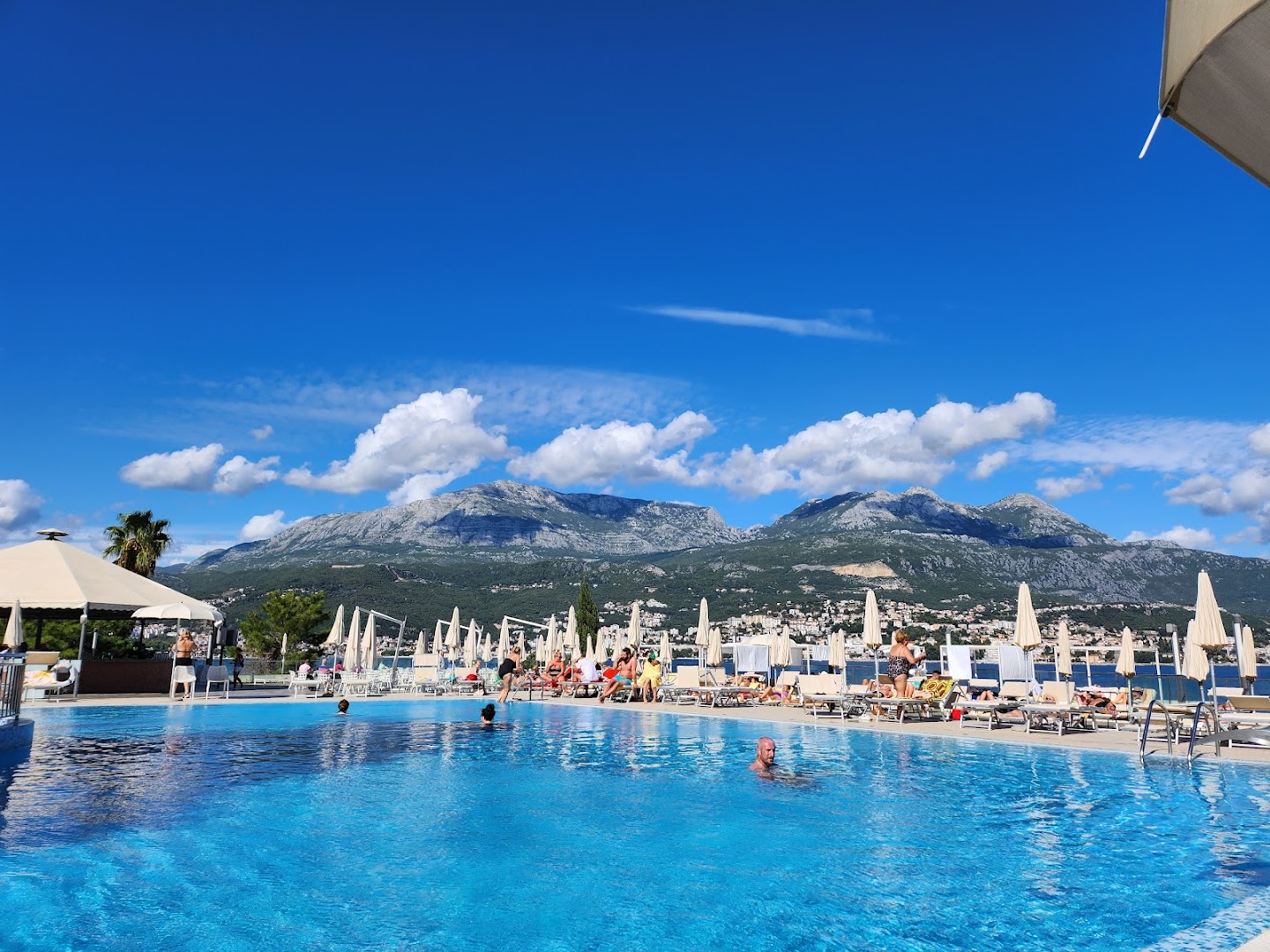 Iberostar Herceg Novi viesi bauda atpūtu lielajā āra baseinā ar skatu uz kalnu grēdām un pilsētas panorāmu tālumā