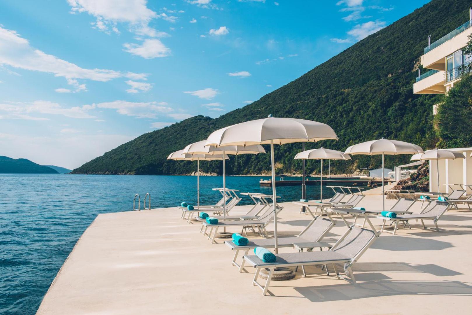 Iberostar Herceg Novi sauļošanās terase pie ūdens — balti saulessargi un krēsli uz betona platformas pie jūras