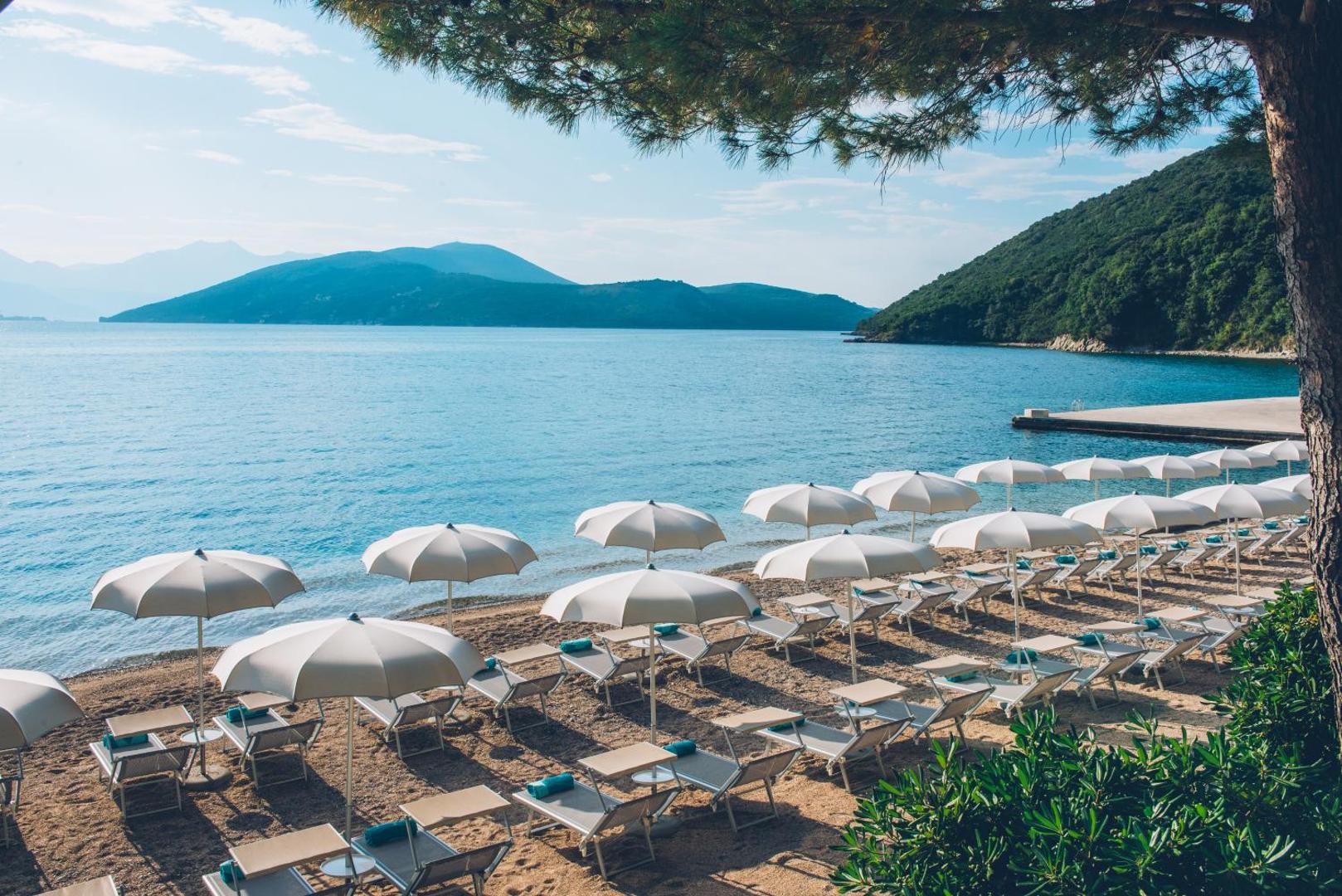 Iberostar Herceg Novi privātā oļu pludmale Melnkalnē — sauļošanās krēsli ar saulessargiem līča krastā un kalnu fons