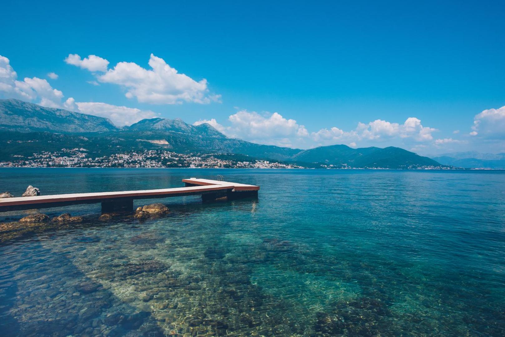 Iberostar Herceg Novi koka laipa kristāldzidrā Adrijas jūrā Melnkalnē ar skatu uz kalniem un piekrastes pilsētu tālumā