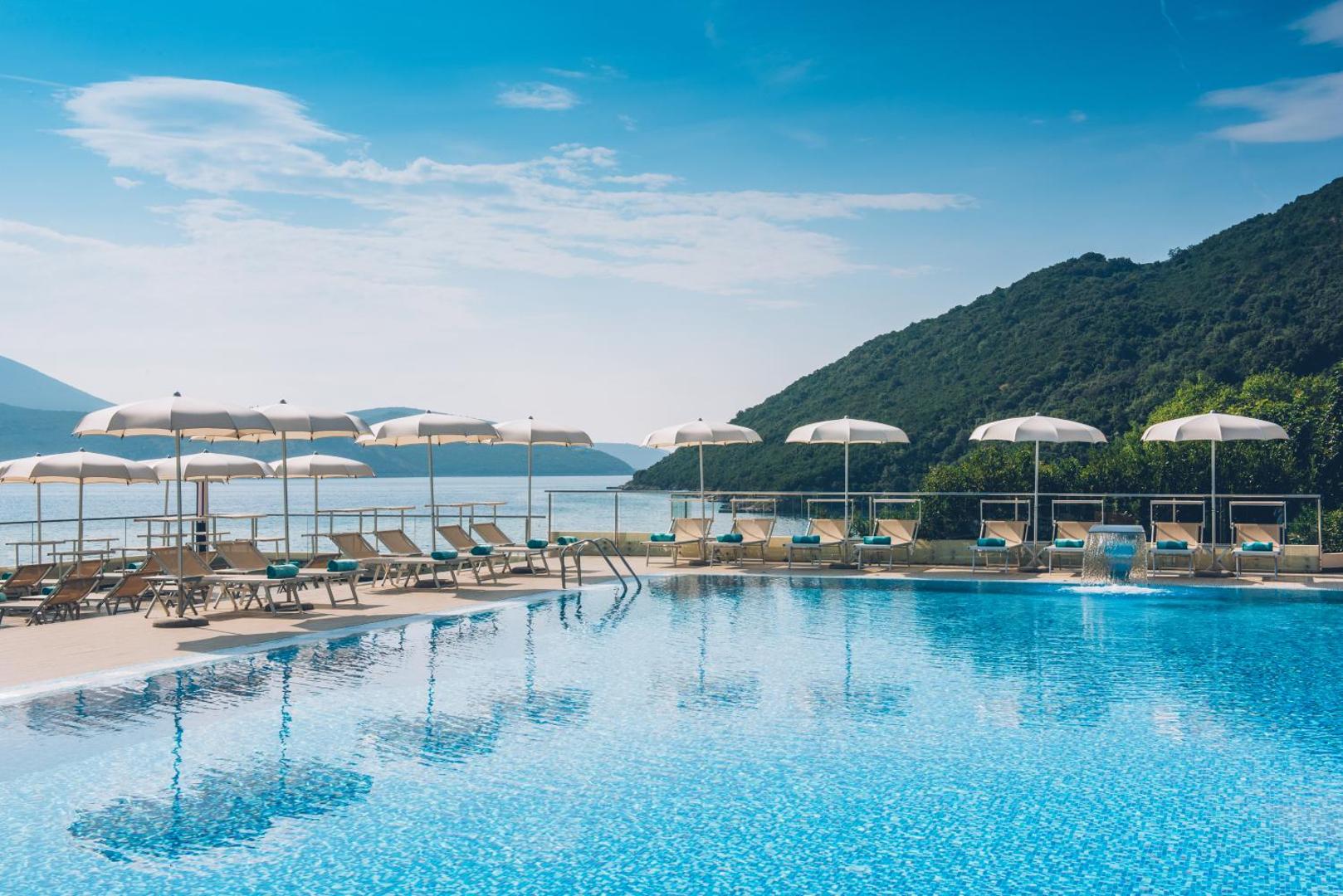 Viesnīcas Iberostar Herceg Novi baseins un sauļošanās terase kalnu pakājē — gaiši saulessargi un zili dvieļi uz zviļņiem