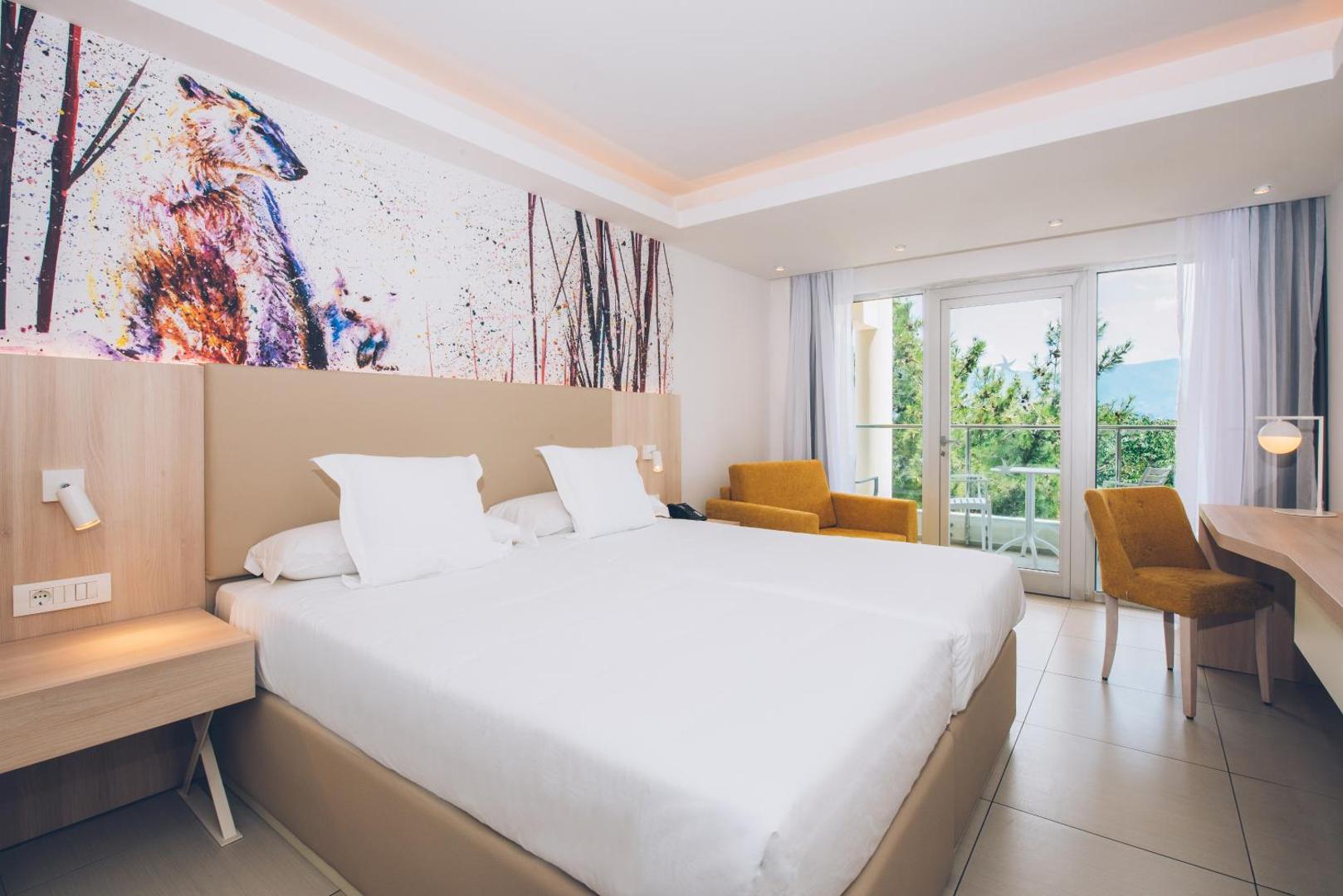Iberostar Herceg Novi moderns viesnīcas numurs ar lielu gultu, māksliniecisku sienas dekoru un skatu uz dārzu caur balkona durvīm