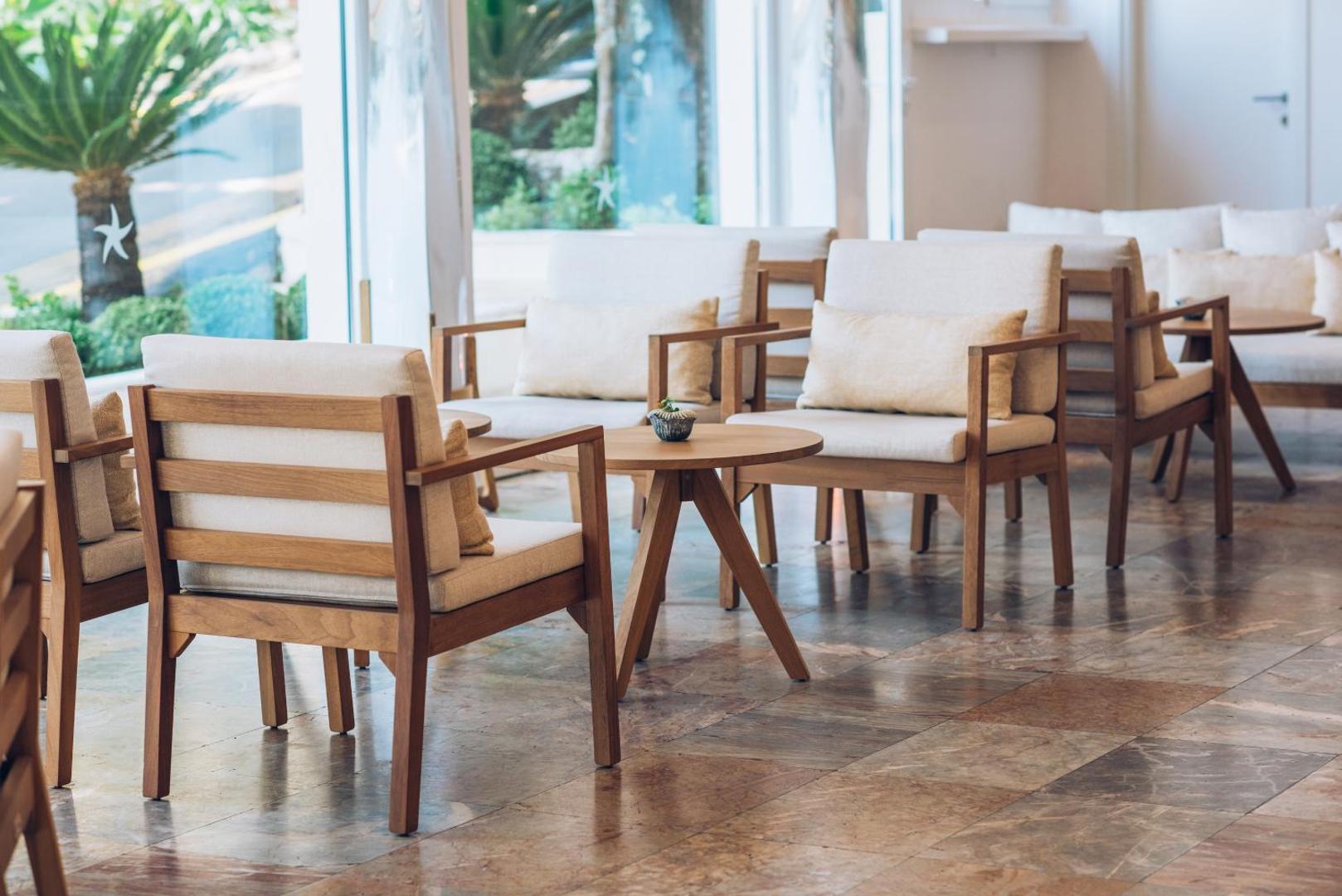 Iberostar Herceg Novi viesnīcas lobby atpūtas zona ar koka krēsliem, gaišiem spilveniem un skatu uz dārzu caur lielajiem logiem