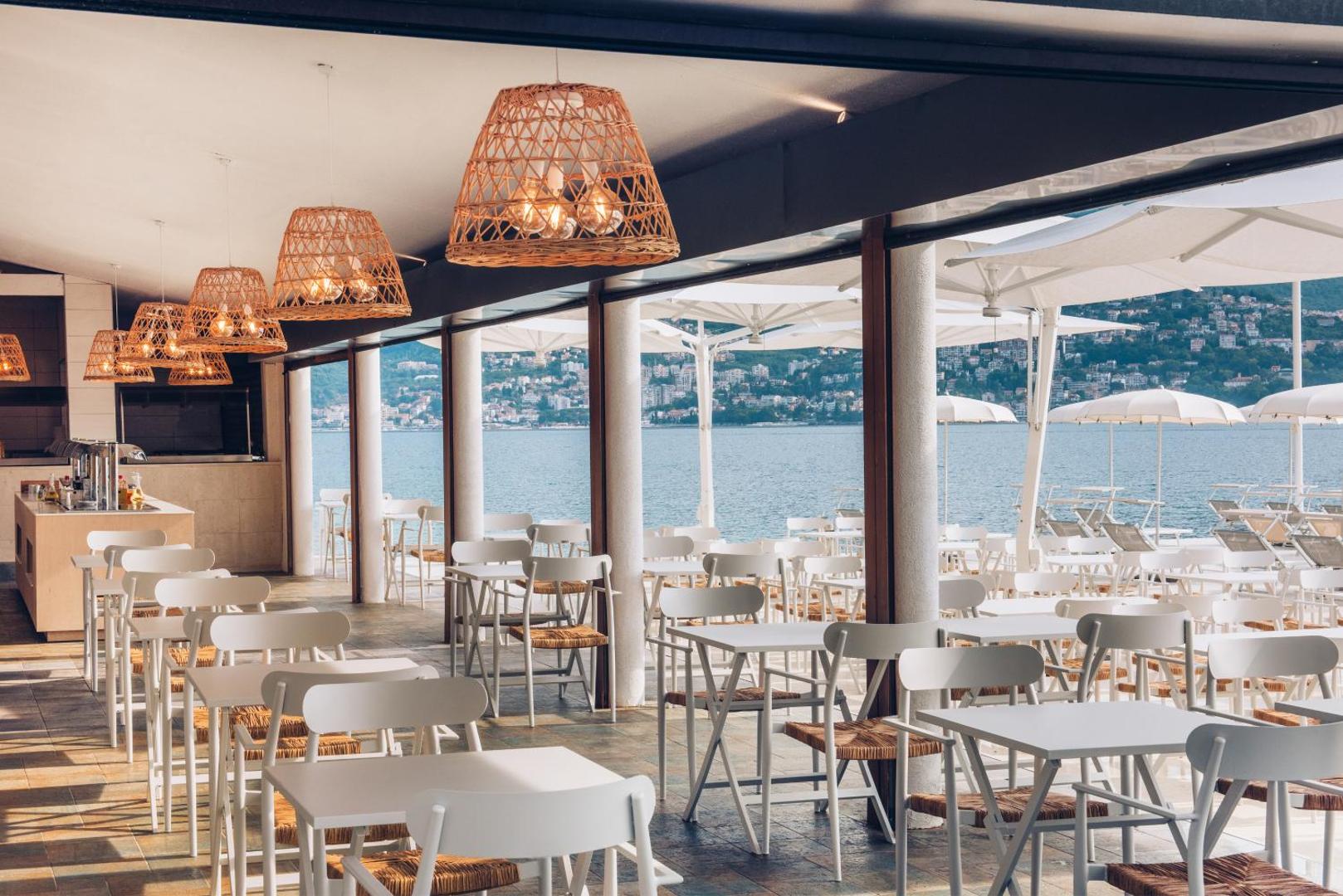 Iberostar Herceg Novi viesnīcas restorāns ar skatu uz jūru — gaišs interjers, pītas lampas un balti galdiņi piekrastē