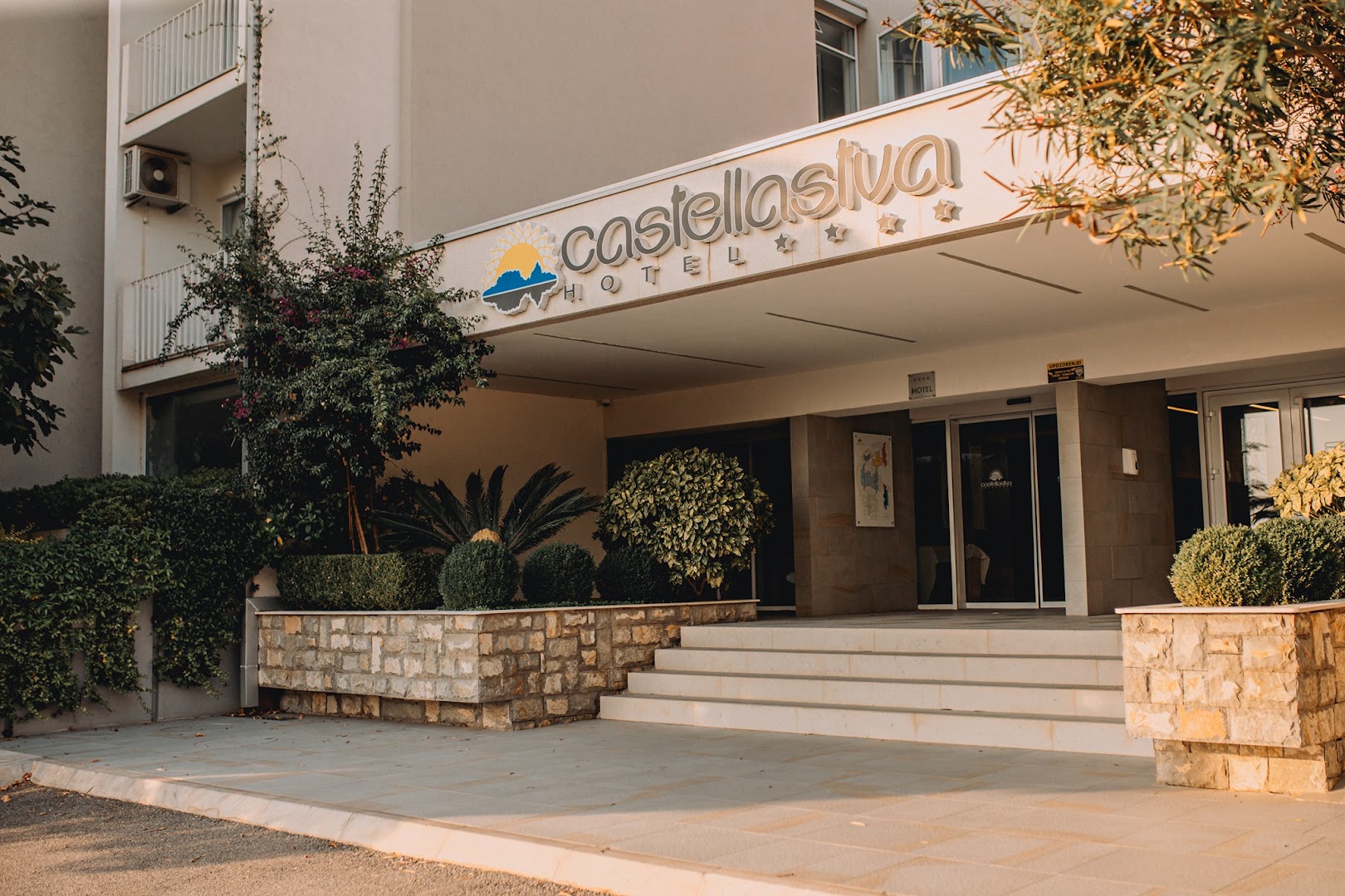 Tuvplāns Hotel Castellastva ieejas zonai ar viesnīcas nosaukumu, akmens mūrējumu un dekoratīviem krūmiem pēcpusdienas saulē