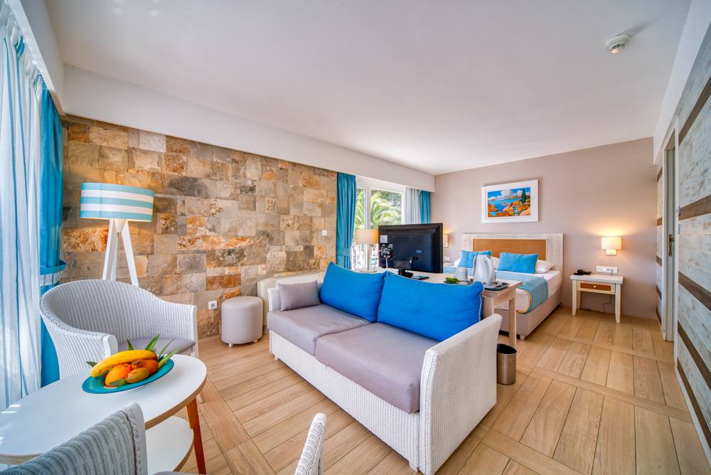 Hotel Castellastva numura dzīvojamā zona ar dīvānu, atpūtas krēsliem un TV Petrovacā — gaišs interjers ar koka un akmens elementiem