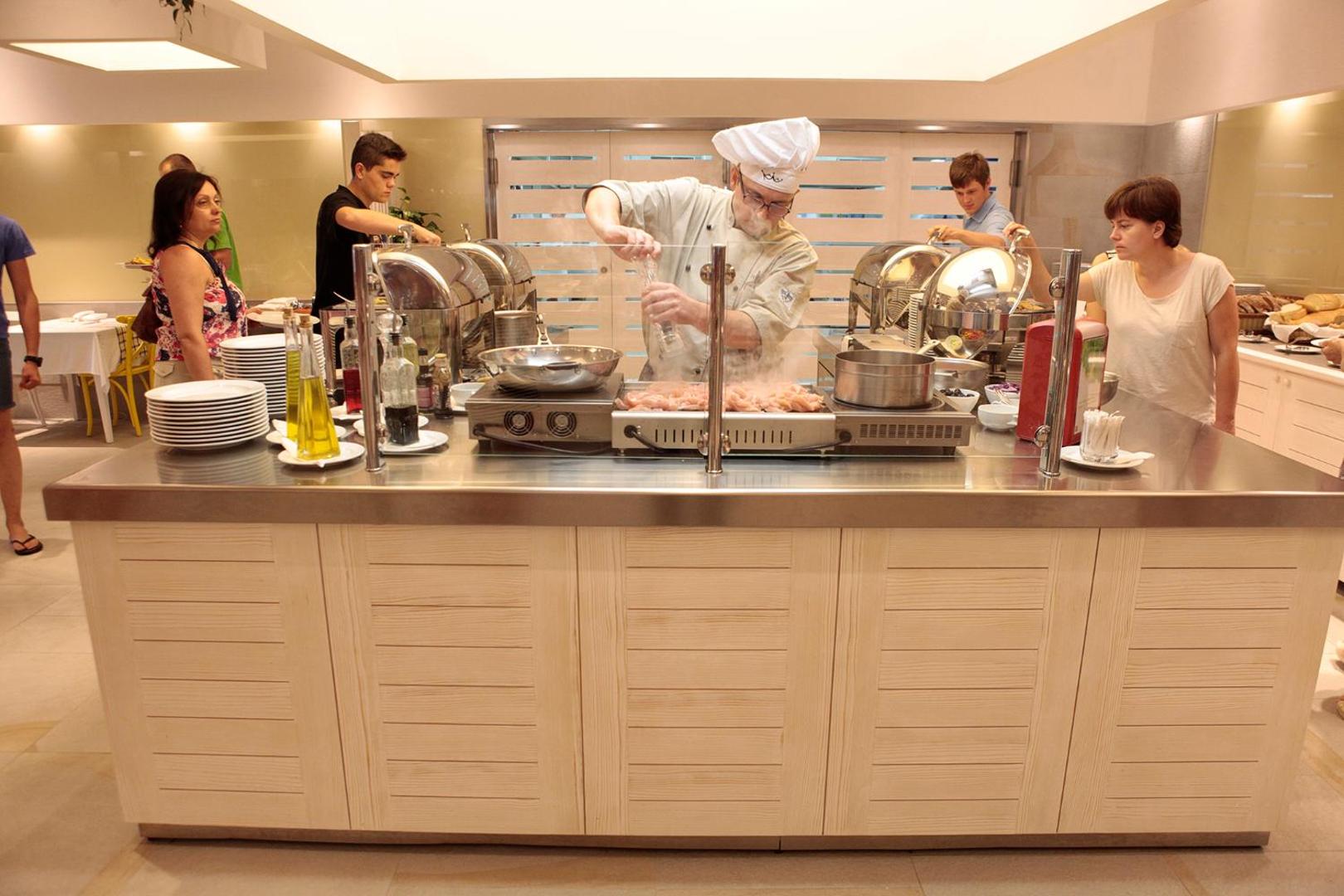 Šefpavārs gatavo ēdienu Hotel Castellastva atvērtā tipa virtuvē — bufetes zona ar siltajiem ēdieniem un svaigiem produktiem
