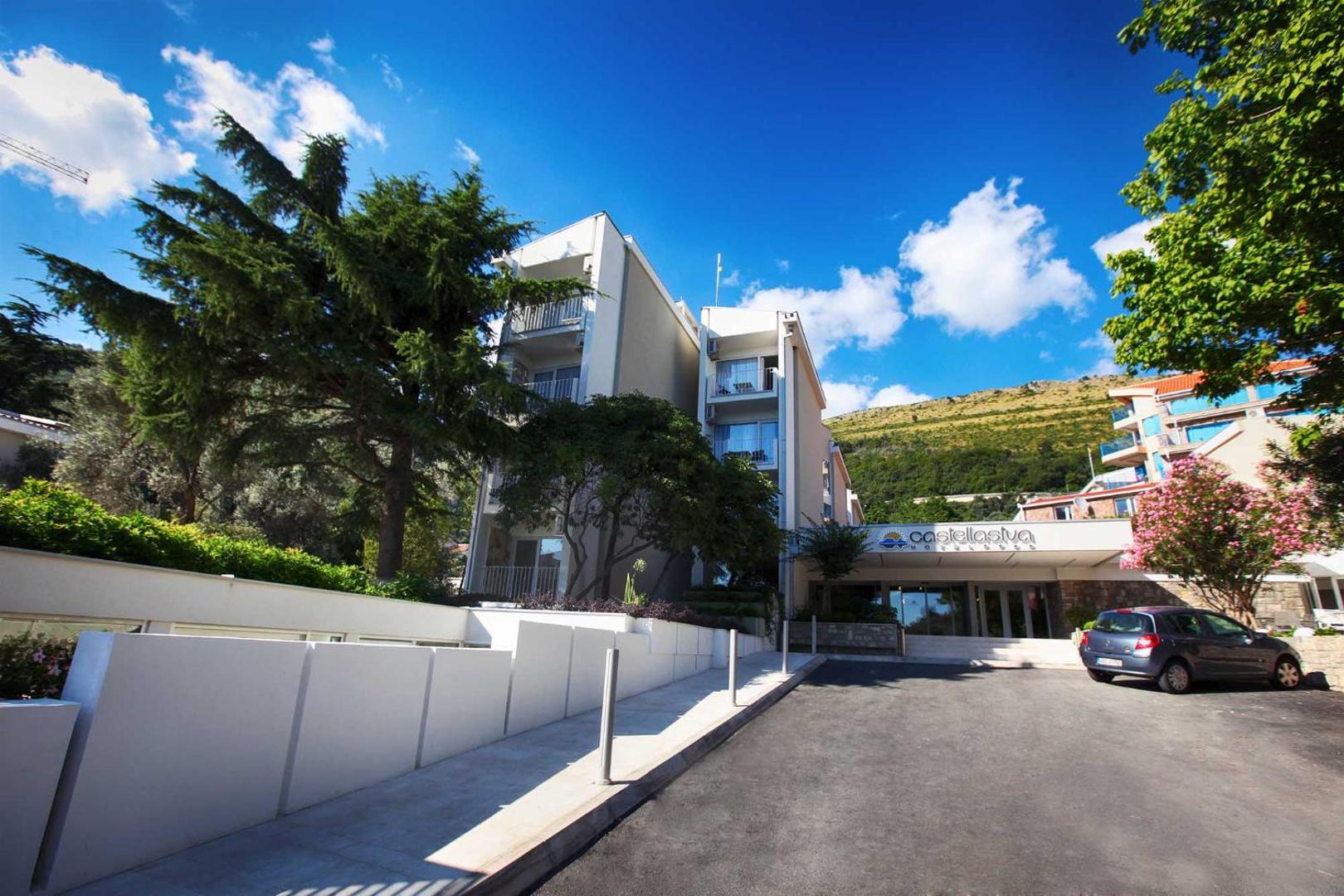 Hotel Castellastva ēkas fasāde ar balkoniem uz kalnu fona Petrovacā, Melnkalnē — plašs skats uz teritoriju un piebraucamo ceļu
