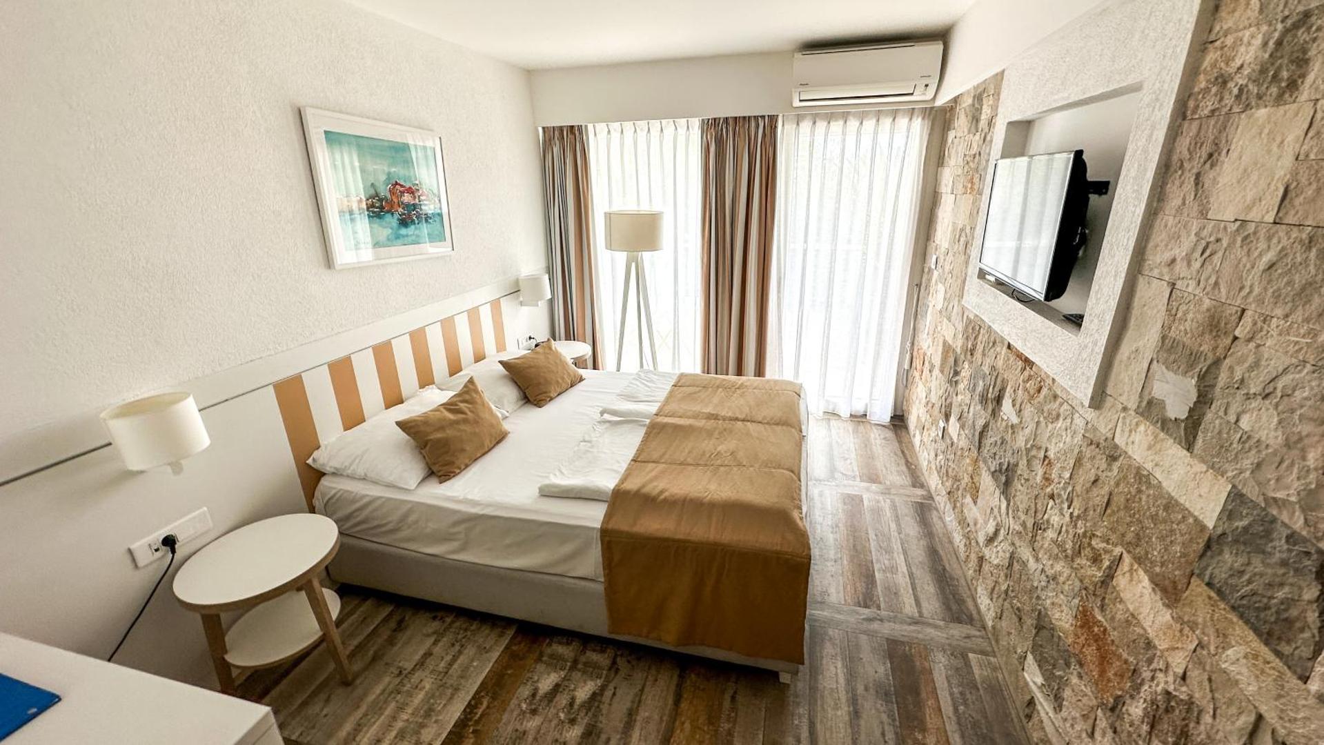 Hotel Castellastva divvietīgs numurs ar smilšu krāsas tekstilu un akmens sienas dekoru Petrovacā — lieli logi nodrošina dabisku gaismu