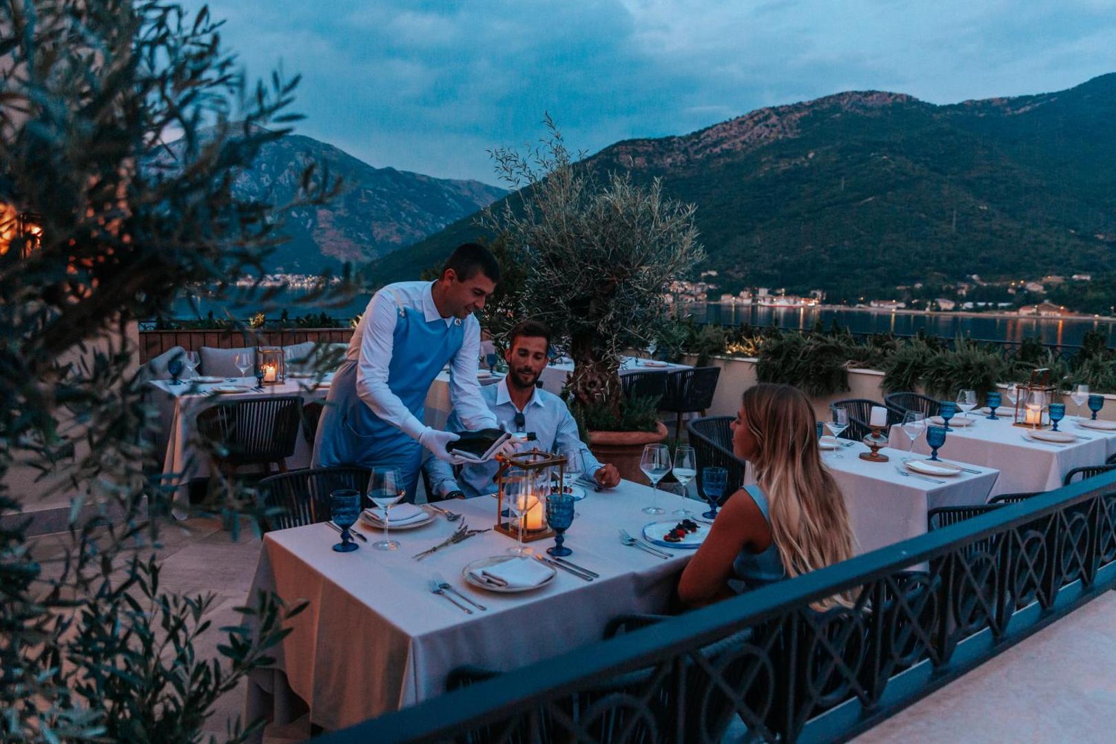 Boutique Hotel & Spa Casa del Mare restorāna terase vakarā ar skatu uz kalniem un jūru, viesmīlis apkalpo pāri pie galdiņa