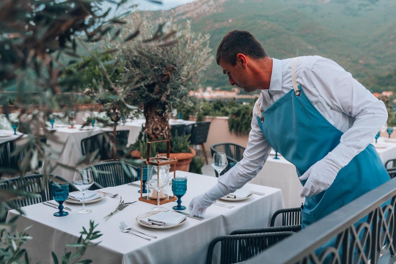 Viesnīcas darbinieks klāj galdu uz Boutique Hotel & Spa Casa del Mare āra terases, fonā redzami olīvkoki un kalni
