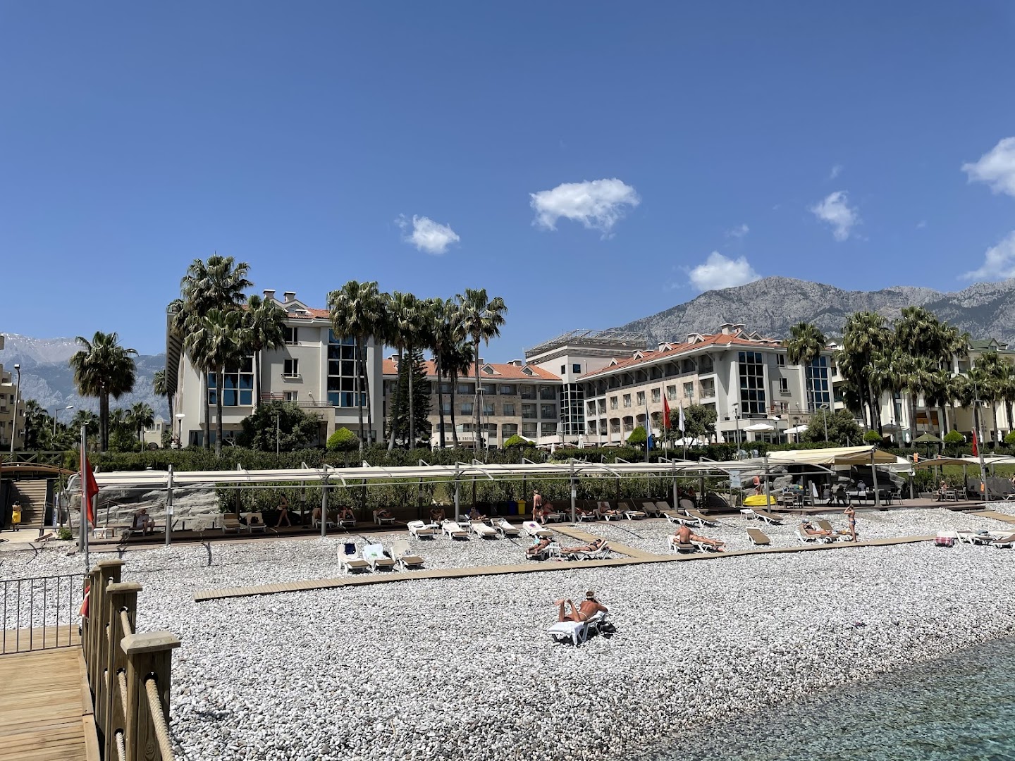 Fame Residence Kemer & Spa viesnīcas ēka un plašā oļu pludmale ar saulessargiem un atpūtas krēsliem uz kalnu fona
