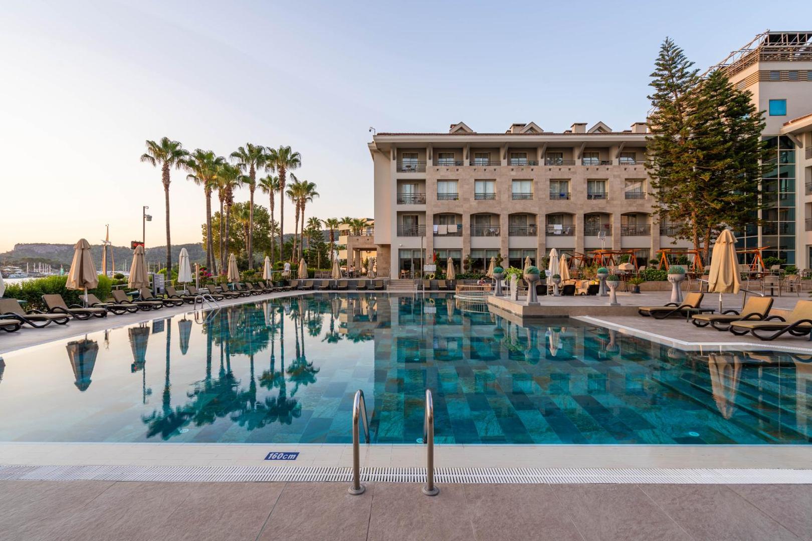 Fame Residence Kemer & Spa āra baseins ar palmu atspulgu ūdenī un viesnīcas numuru balkoniem fonā