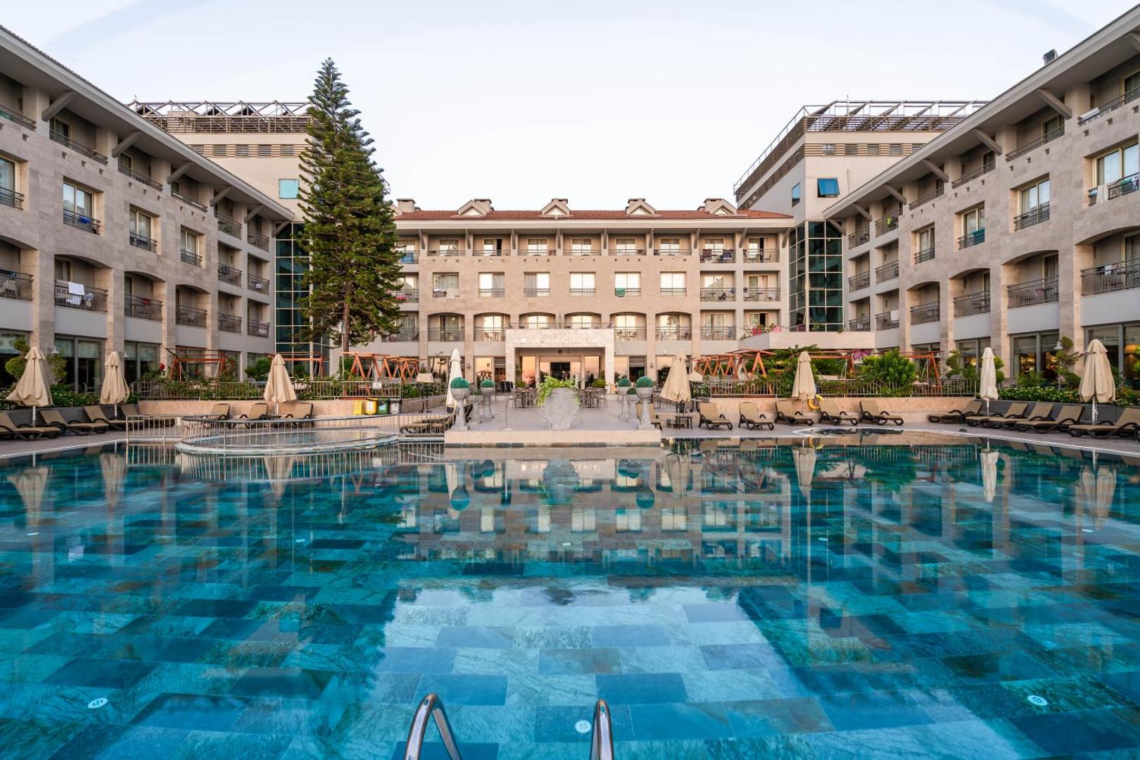 Fame Residence Kemer & Spa galvenais āra baseins un viesnīcas ēkas fasāde vakarā — atpūtas zona ar saulessargiem un krēsliem