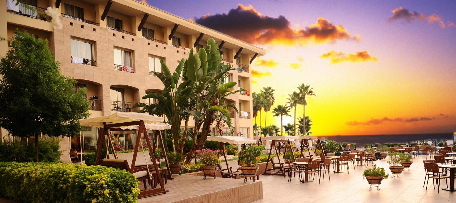 Fame Residence Kemer & Spa viesnīcas fasāde un āra terase ar atpūtas šūpolēm un galdiņiem saulrieta gaismā Kemeras piekrastē
