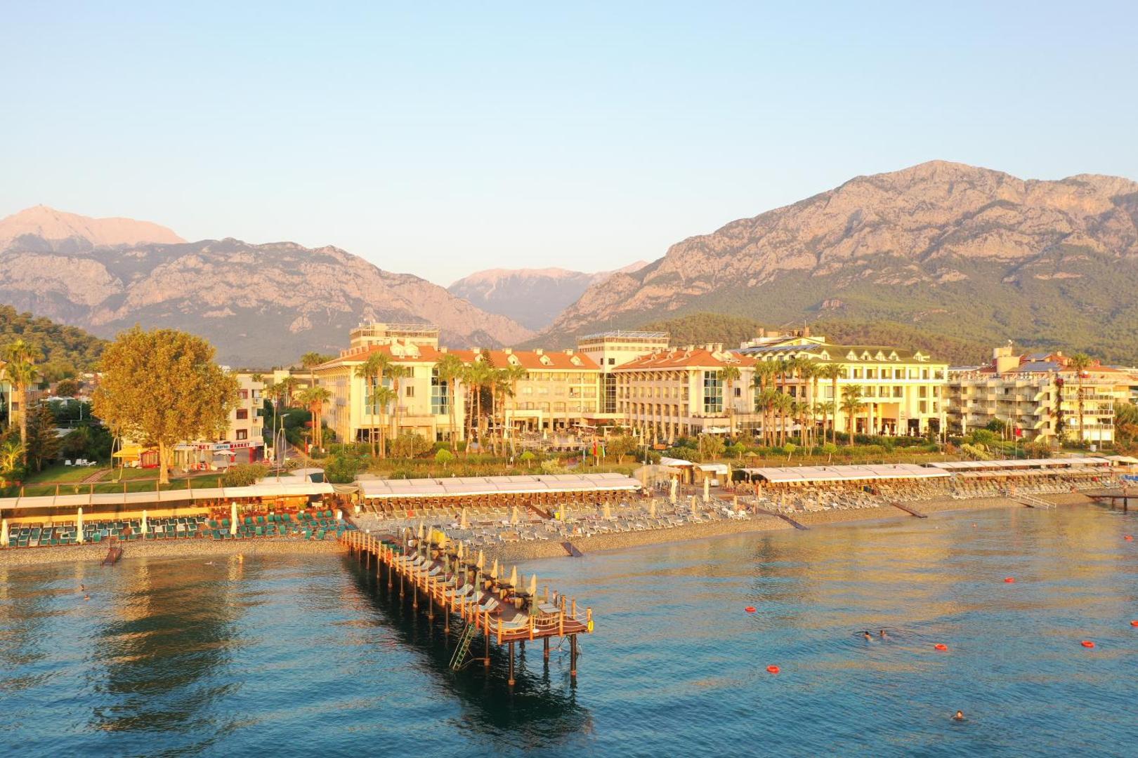 Fame Residence Kemer & Spa skats no jūras puses uz viesnīcas ēku, oļu pludmali un majestātiskajiem Tauru kalniem saulrietā