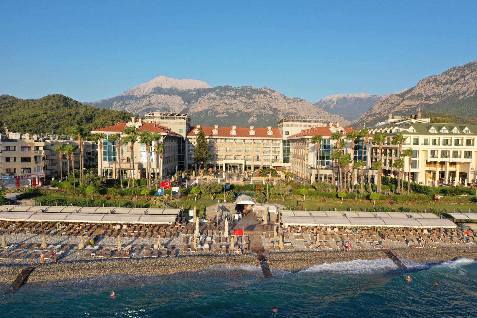Fame Residence Kemer & Spa fasāde un pludmales zona ar saulessargiem, fonā redzamas augstās kalnu grēdas skaidrās debesīs