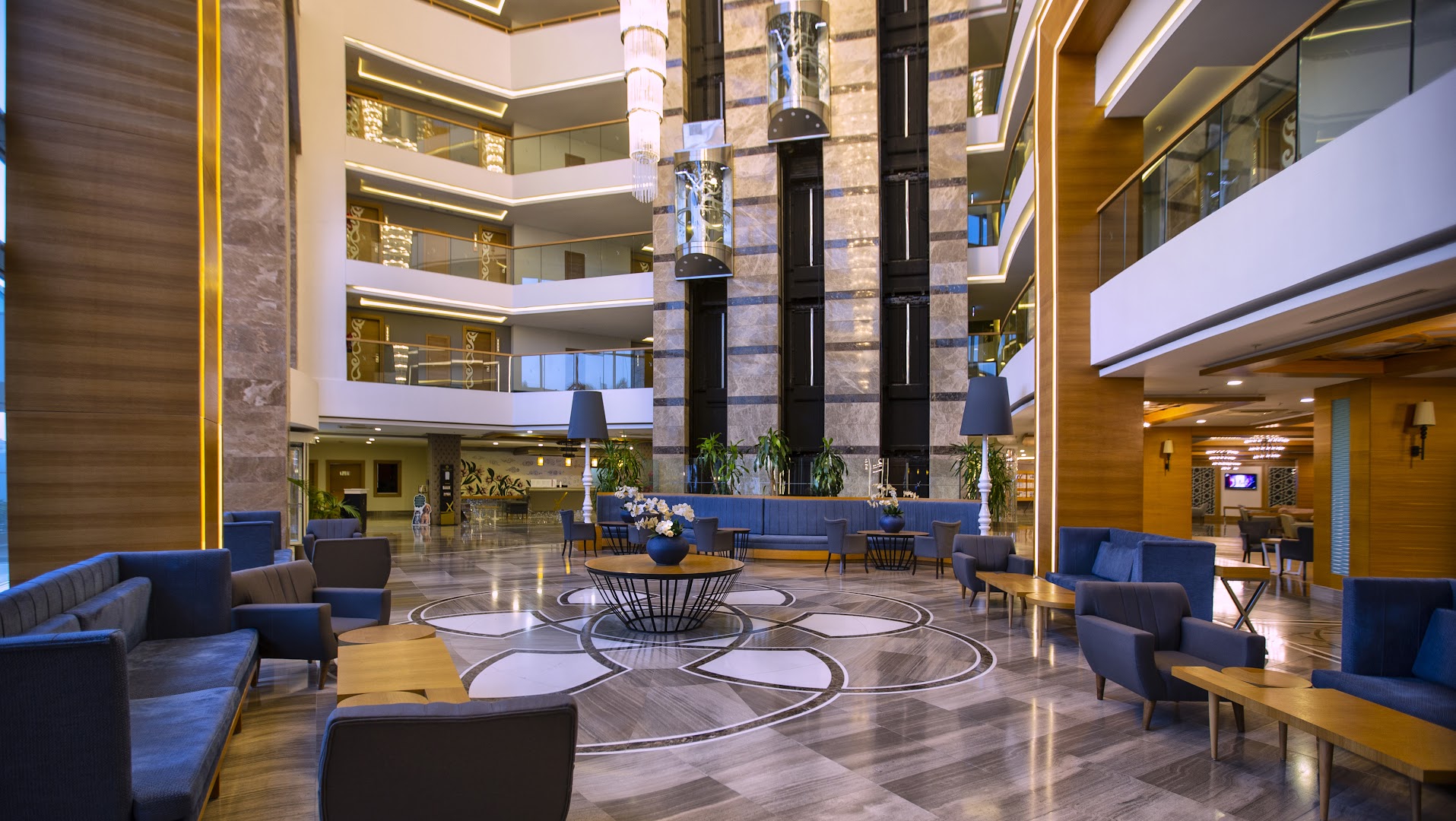 Sherwood Exclusive Lara plašais viesnīcas lobby ar moderniem atpūtas krēsliem, augstiem griestiem, stikla liftiem un greznām lustrām