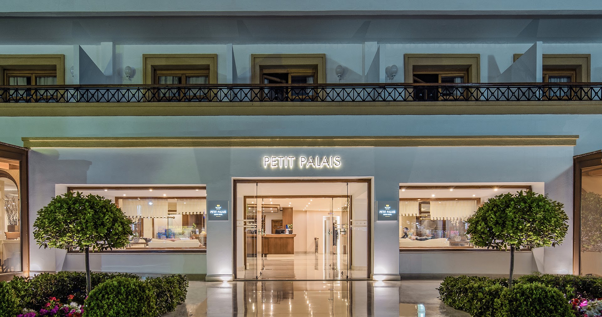 Canvas by Mitsis Petit Palais viesnīcas ieeja vakarā Rodas pilsētā — izgaismota fasāde ar stikla durvīm, koptiem apstādījumiem un lobby zonas skatu