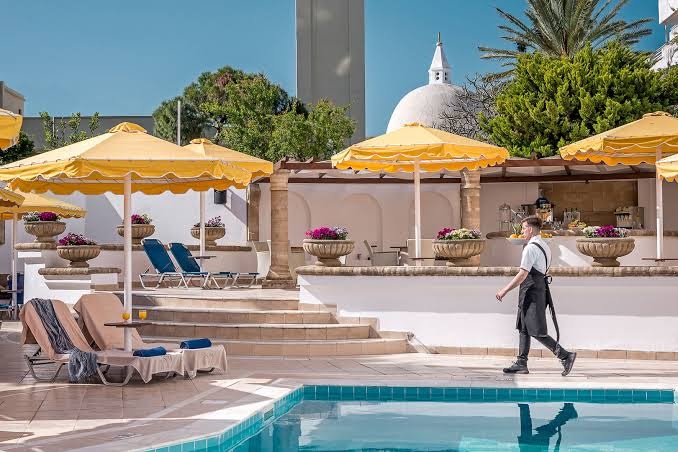 Mitsis Petit Palais viesnīcas baseina terase Rodā ar sauļošanās krēsliem, dzelteniem saulessargiem un skatu uz pilsētas arhitektūru.