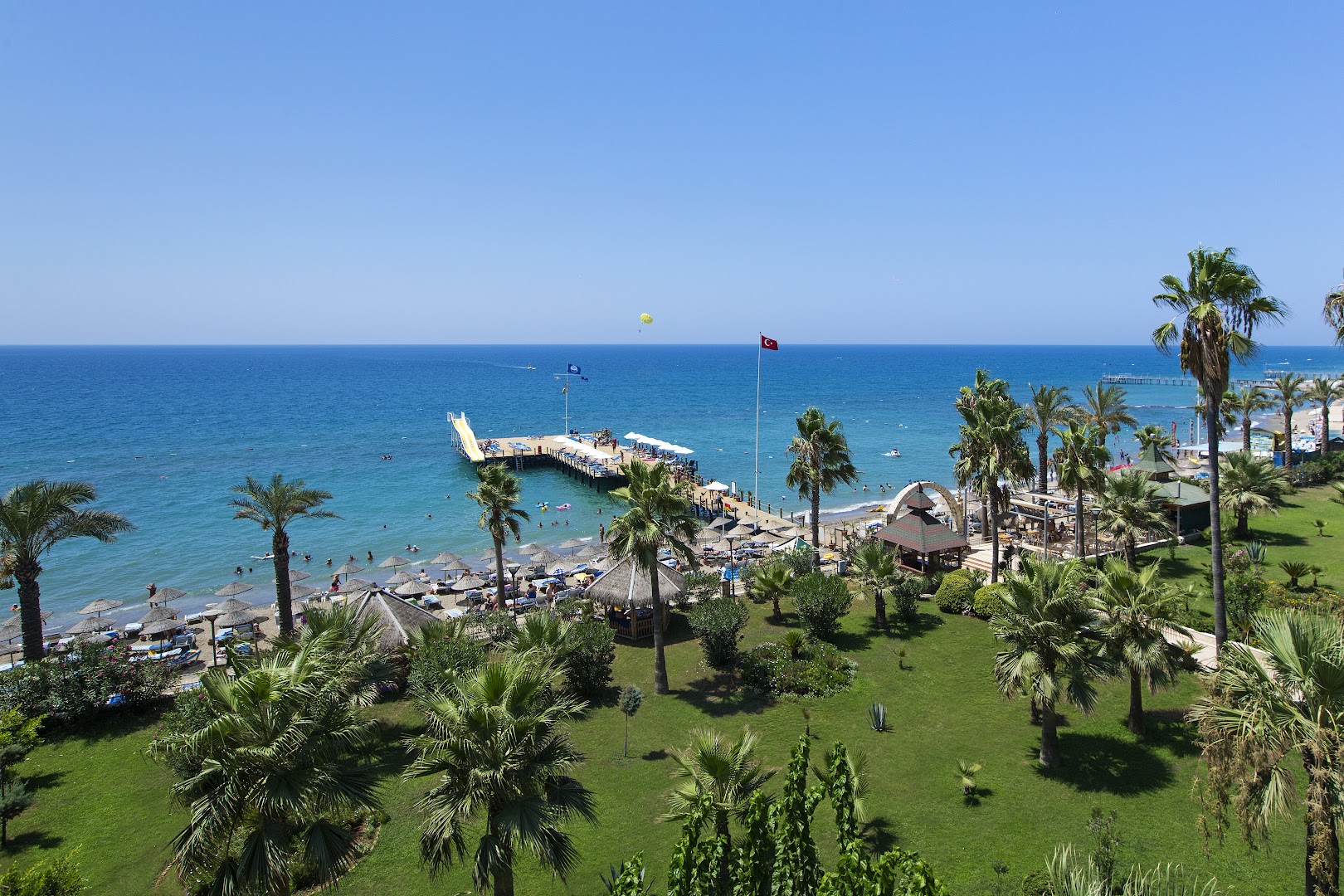Saphir Hotel & Villas skats no augšas uz Vidusjūras piekrasti, zaļo dārzu ar palmām un koka piestātni Alanjas reģionā, Turcijā.