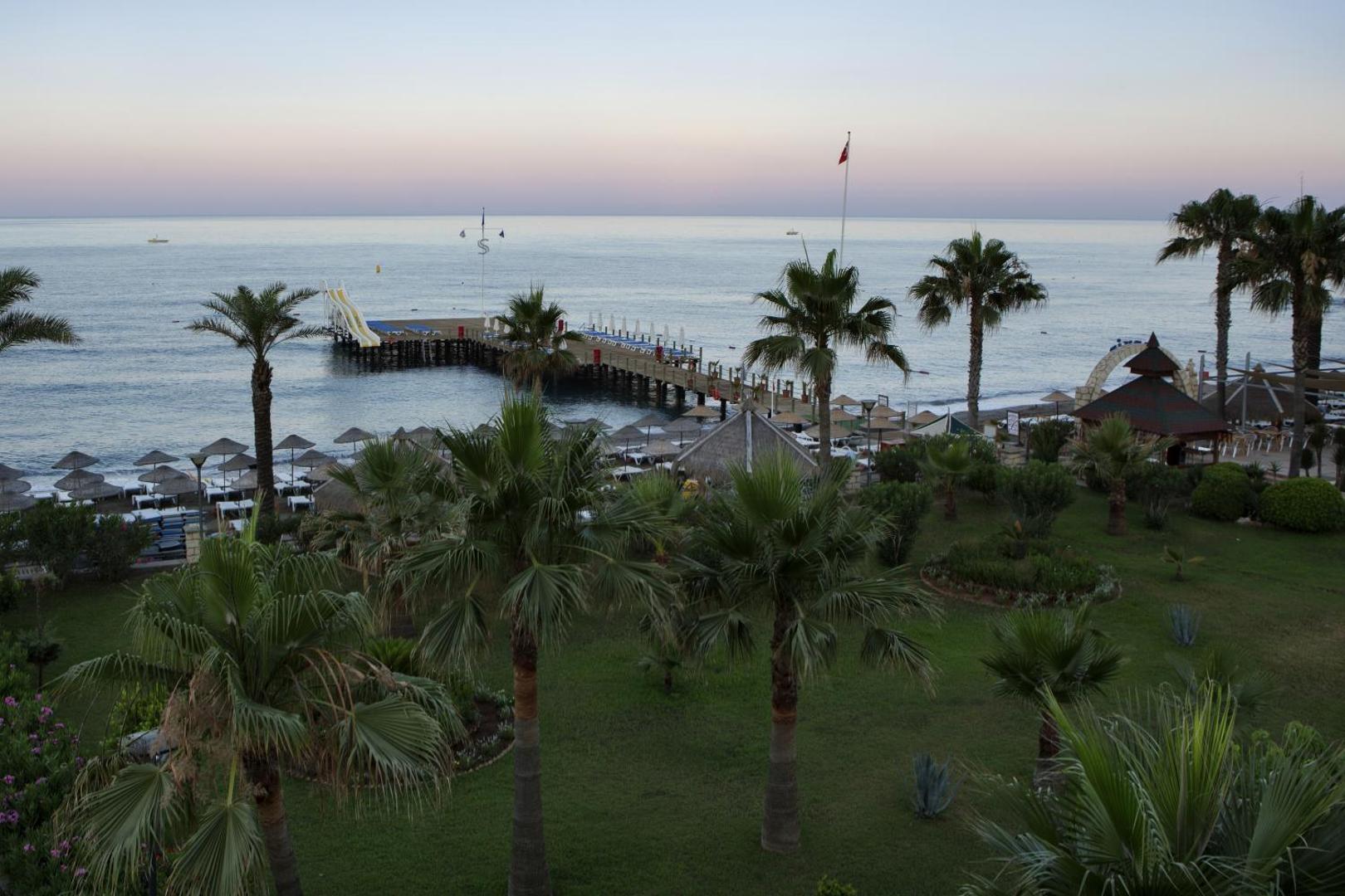 Saphir Hotel & Villas dārzs un piestātne saulrietā — mierīga atmosfēra pie jūras ar palmām un pludmales saulessargiem Alanjā.