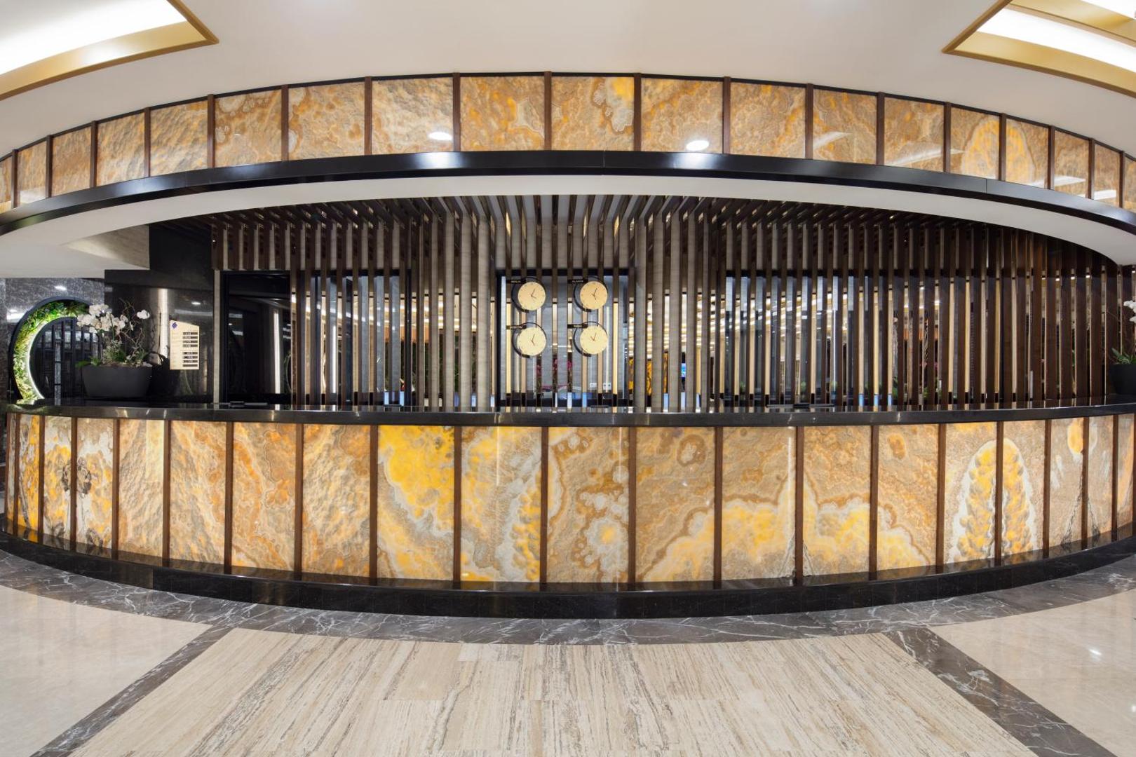 Saphir Hotel & Villas modernā recepcijas zona ar izgaismotu oniksa apdari un pasaules pulksteņiem pie sienas viesnīcas vestibilā.