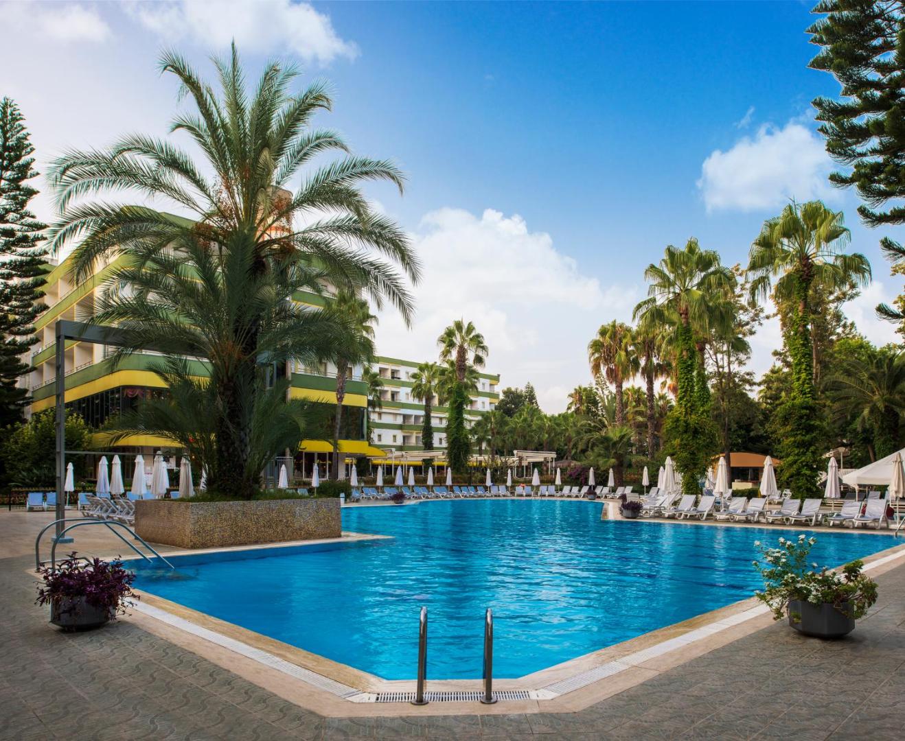 Botanik Hotel & Resort āra baseins ar palmām un sauļošanās krēsliem Alanjas reģionā, Turcijā — skats uz viesnīcas ēku