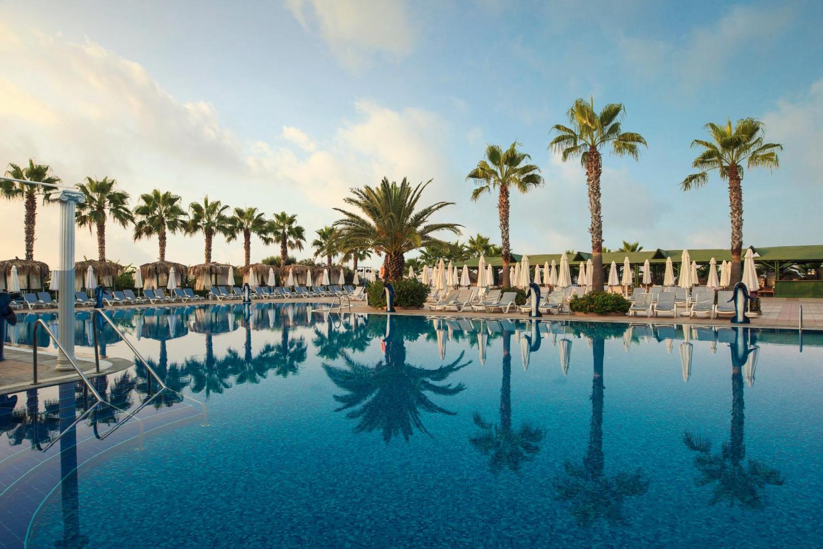 Botanik Hotel & Resort baseina zona saulrietā ar palmām un saulessargiem — ūdens spoguļattēls un mierīga atmosfēra kūrortā