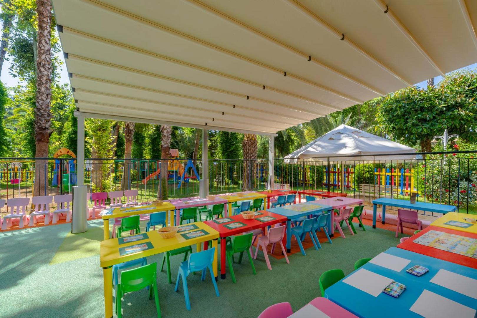 Botanik Hotel & Resort bērnu kluba āra terase ar krāsainiem galdiem un krēsliem radošām nodarbībām zem nojumes dārzā
