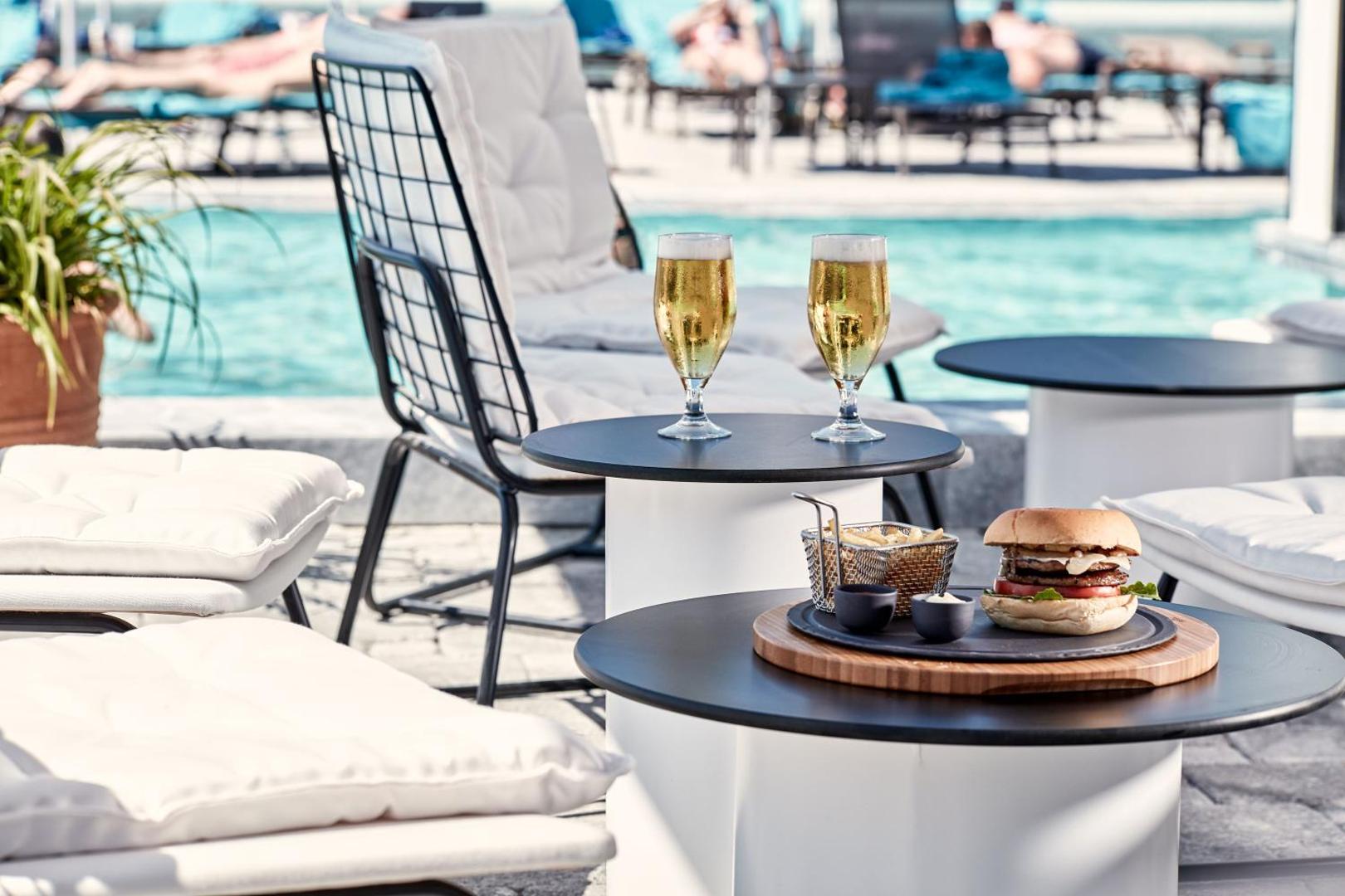 Uzkodas un dzērieni pie Atlantica Dreams Resort baseina — gards burgers un auksts alus uz galdiņa blakus ērtiem sauļošanās krēsliem