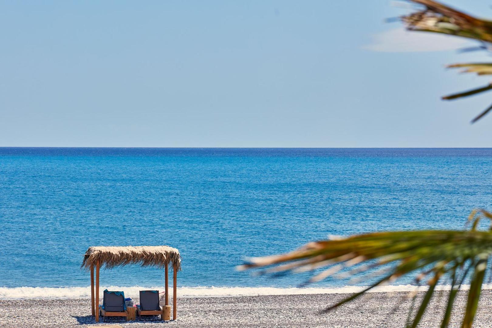 Atlantica Dreams Resort privātā pludmale Rodā ar saulessargu no niedrēm, sauļošanās krēsliem un skatu uz zilo Vidusjūru.