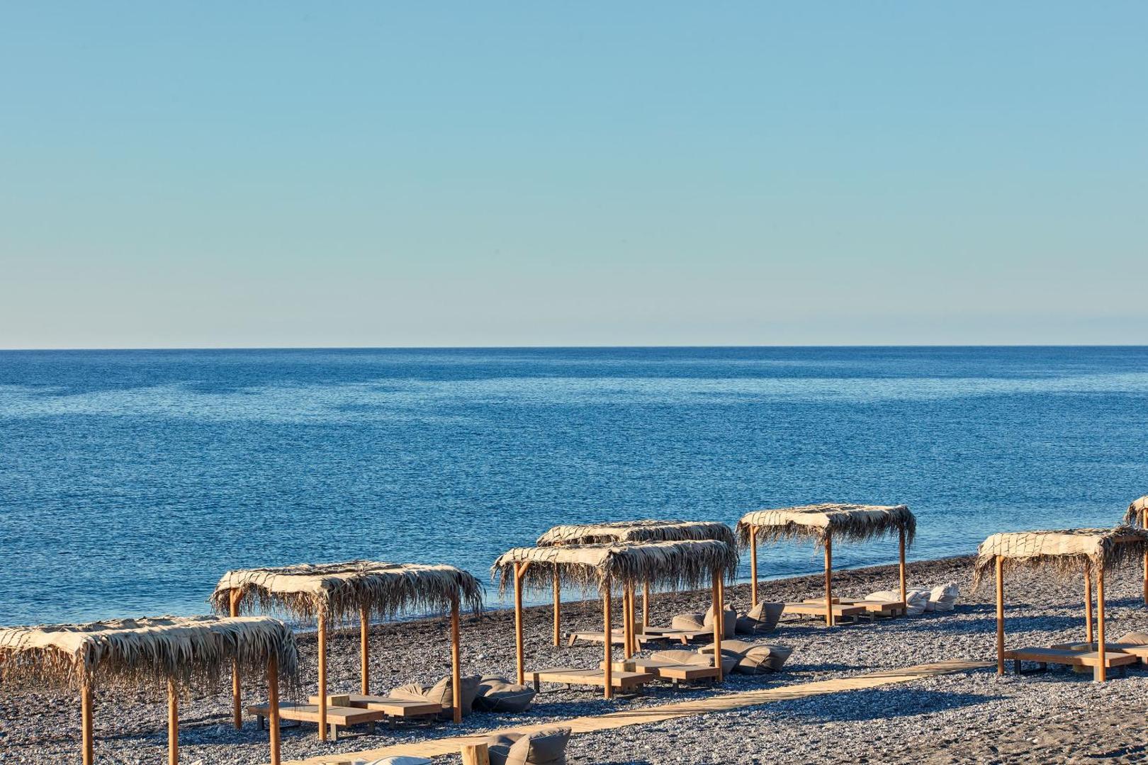 Atlantica Dreams Resort privātā oļu pludmale Rodas salā ar stilīgām niedrēm klātām saulessargu nojumēm un ērtiem zviļņiem pie zilas jūras
