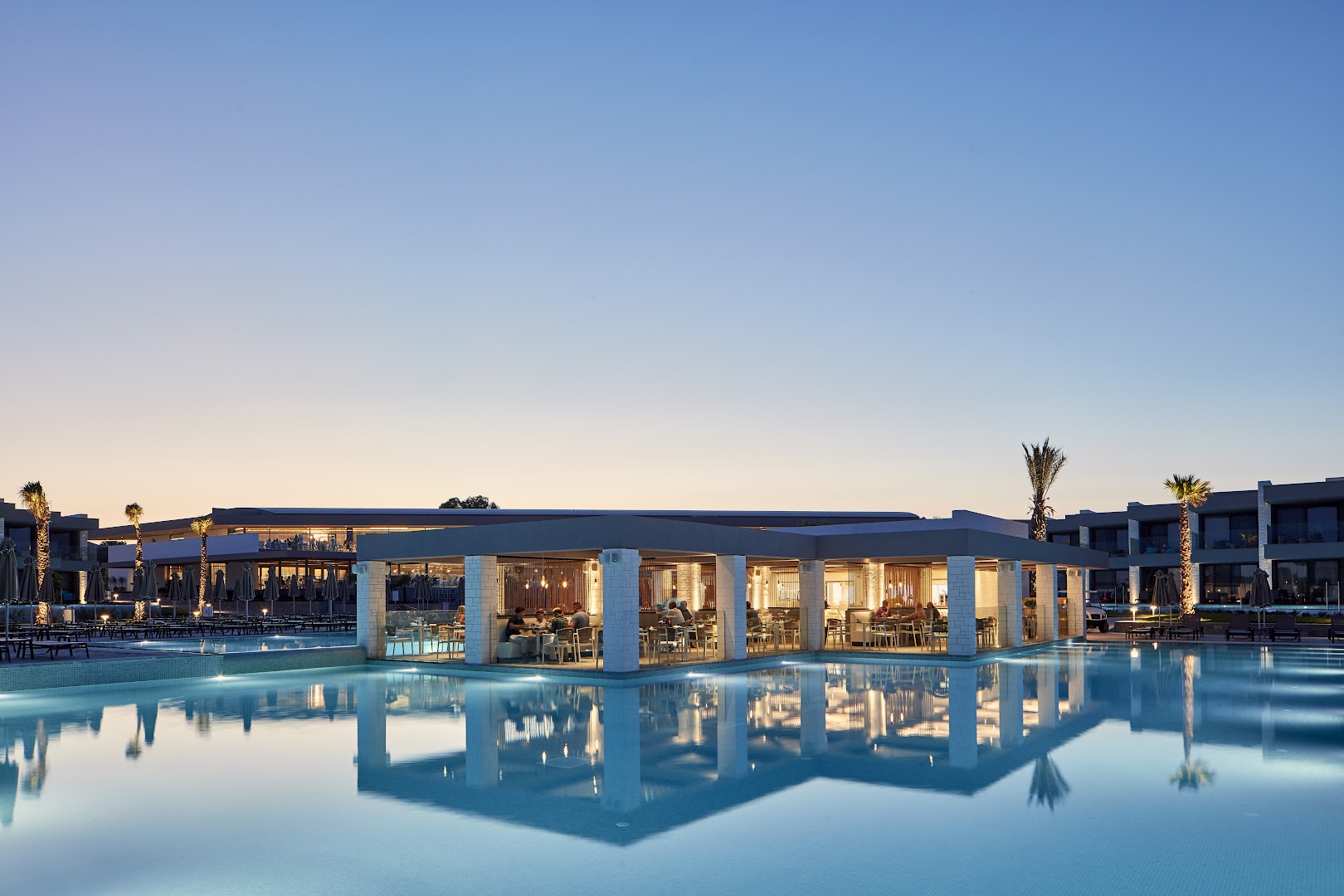 Atlantica Dreams Resort ēdienkartes restorāns vakarā pie izgaismota baseina, moderna arhitektūra un atpūtas zona Rodas piekrastē
