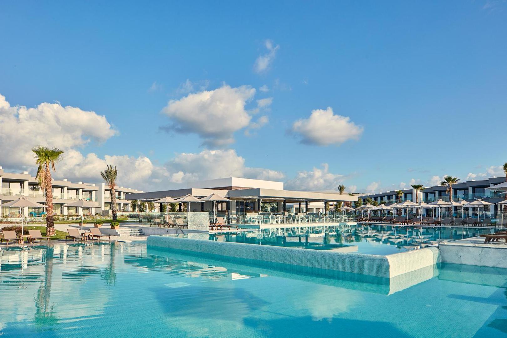 Atlantica Dreams Resort galvenais āra baseins ar pārplūdes malu un saulessargiem Rodā — skats uz modernu restorāna ēku
