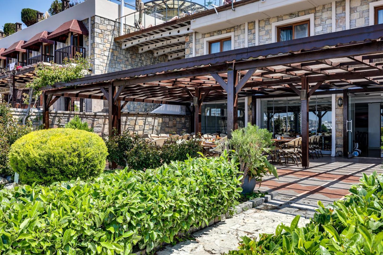 Diamond of Bodrum Hotel āra restorāna zona ar koka pergolu, zaļiem apstādījumiem un akmens mūra fasādi Bodrumā