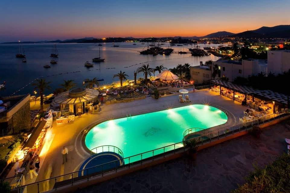 Panorāmas skats no Diamond of Bodrum Hotel terases uz izgaismotu baseinu un saulrietu virs Bodrumas ostas un kalniem.